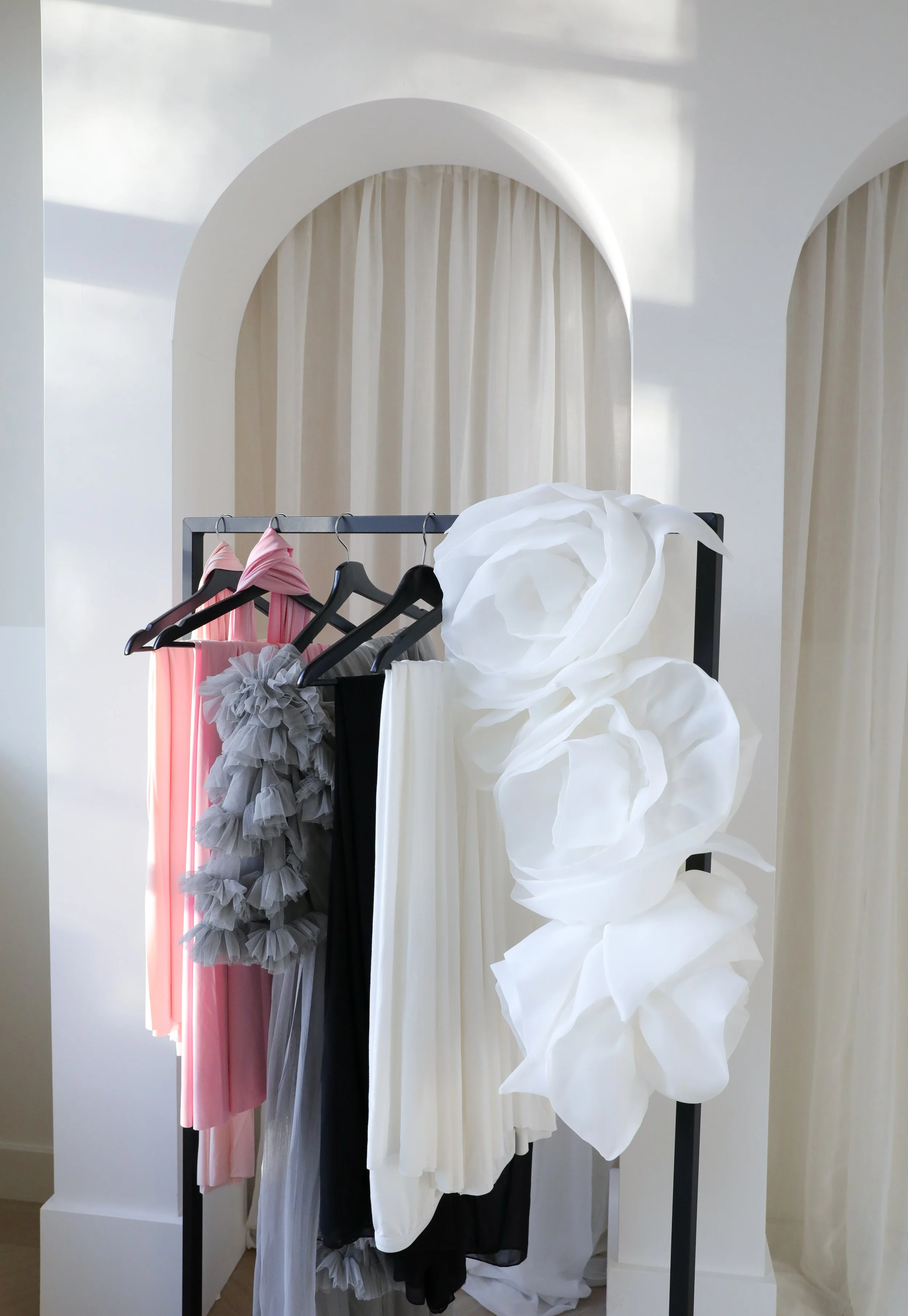 A clothing rack with pink, gray, black, and white dresses, some with ruffles or tulle, in front of white curtains and a wall with archways.