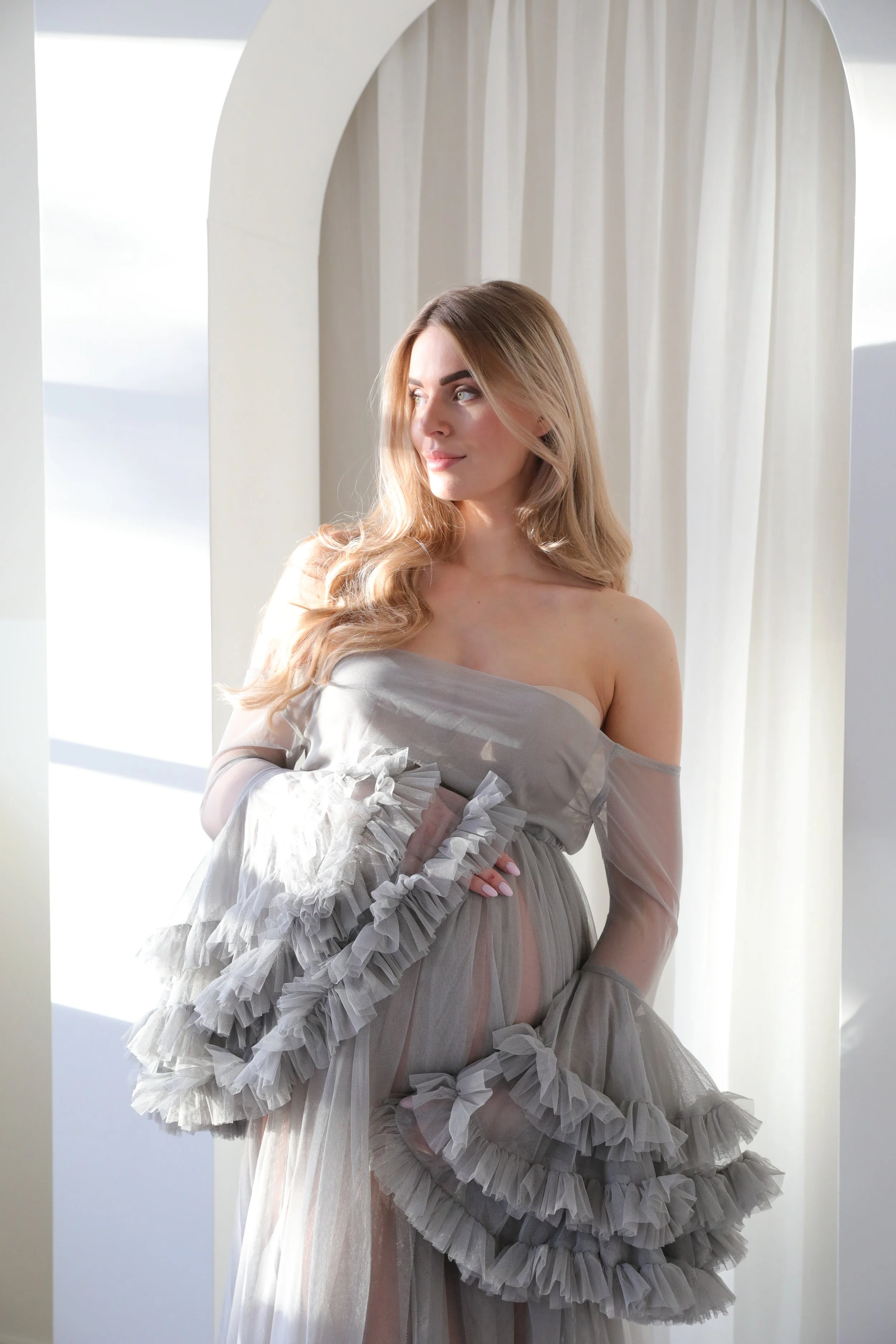 A woman with long blonde hair standing near a white archway with light sheer curtains, wearing an off-shoulder gray dress with ruffled sleeves.