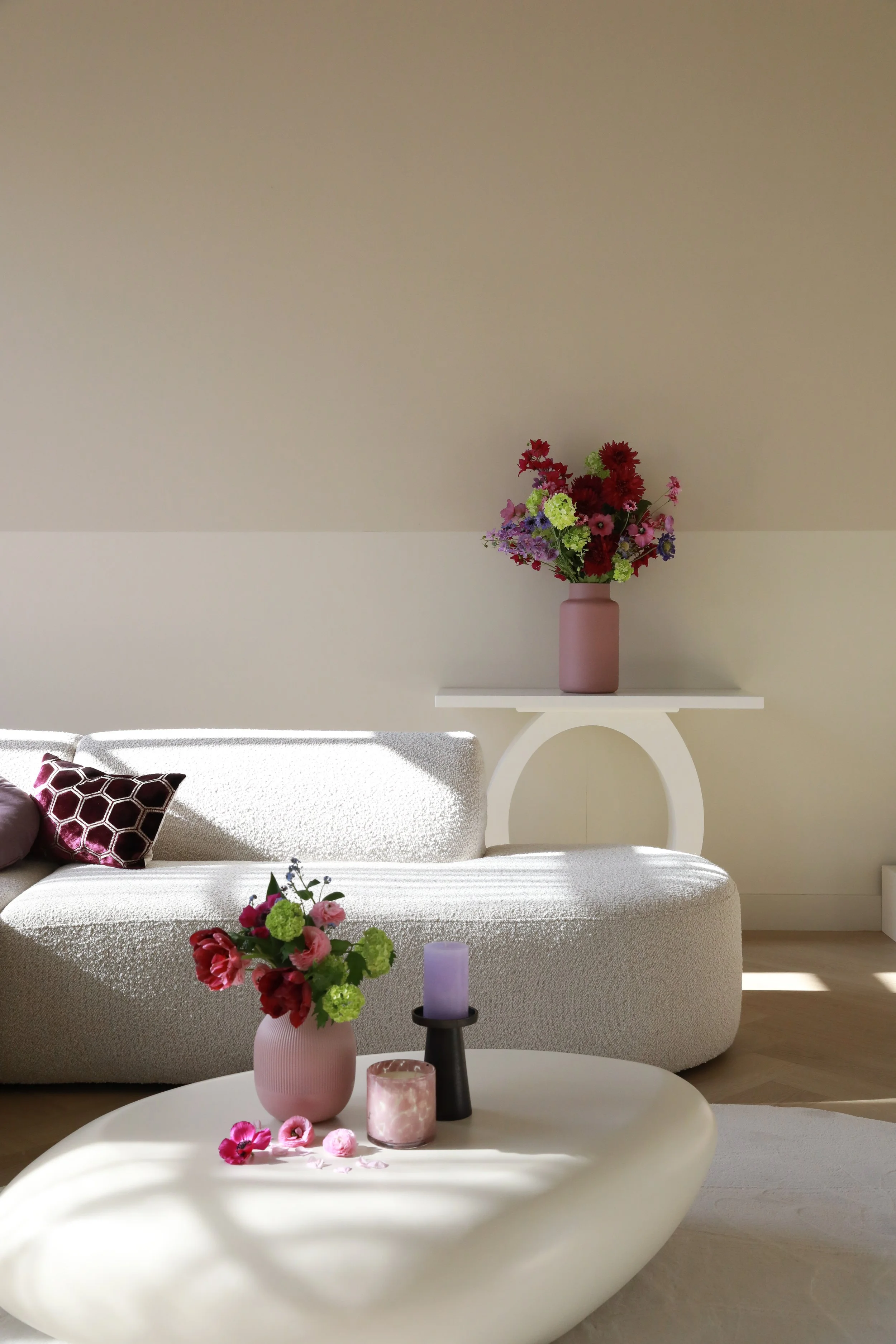 Living room with a white curved sofa, decorated with a pink pillow, and floral arrangements in pink and purple vases. A large vase of flowers is on a white table, and smaller vases with flowers and candles are on the table in the foreground. The background features a wall with a white shelf and another large purple vase with flowers.