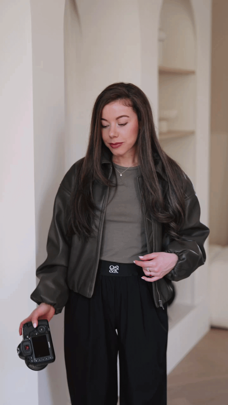 Woman with long brown hair holding a camera, wearing a dark leather jacket and black pants, standing indoors.