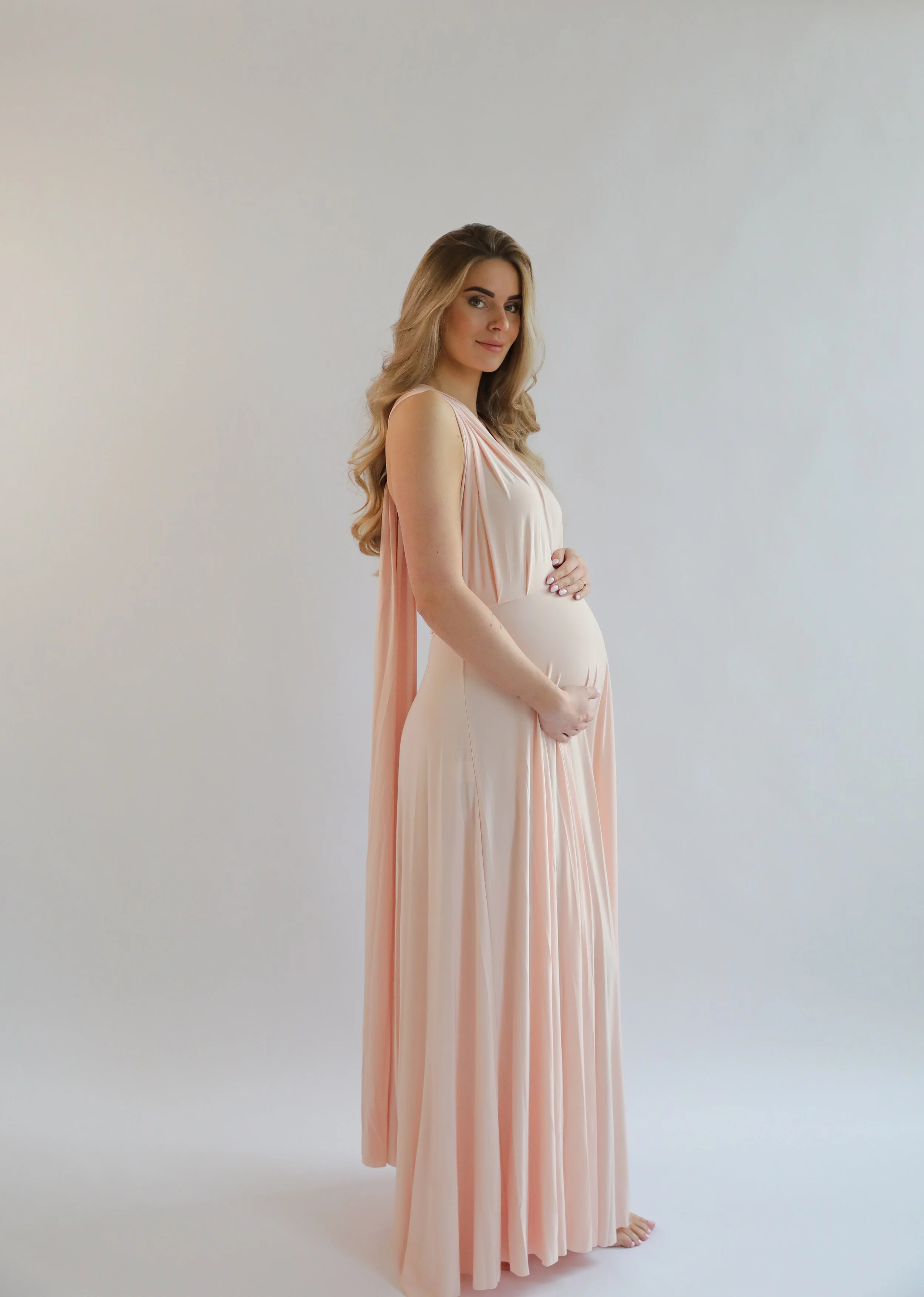 A pregnant woman with long wavy blonde hair in a soft pink and white dress standing barefoot against a plain white background, gently holding her belly with her left hand and smiling softly at the camera.