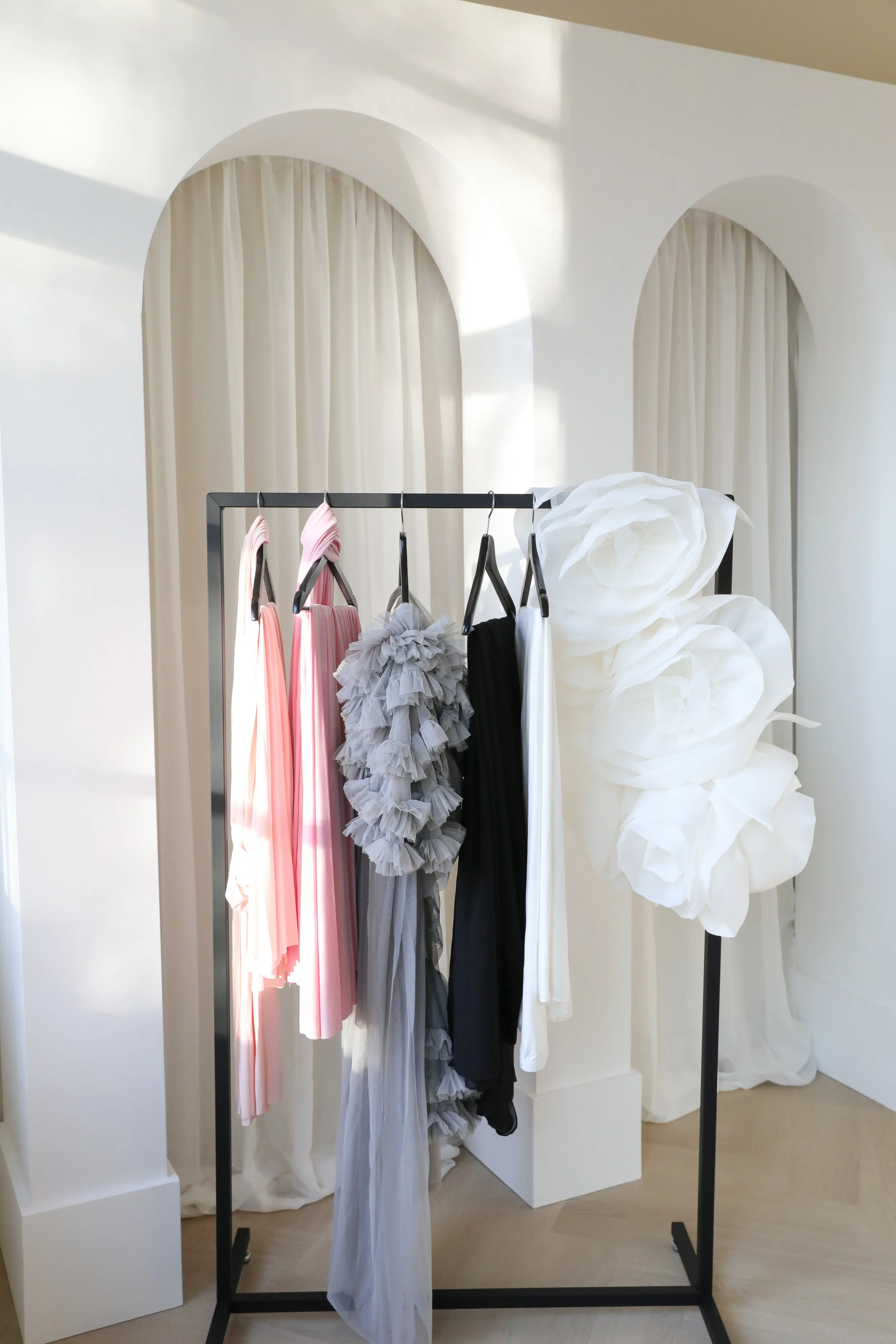 Display of hanging dresses and large white paper flowers in a minimalist room with white walls and curtains.