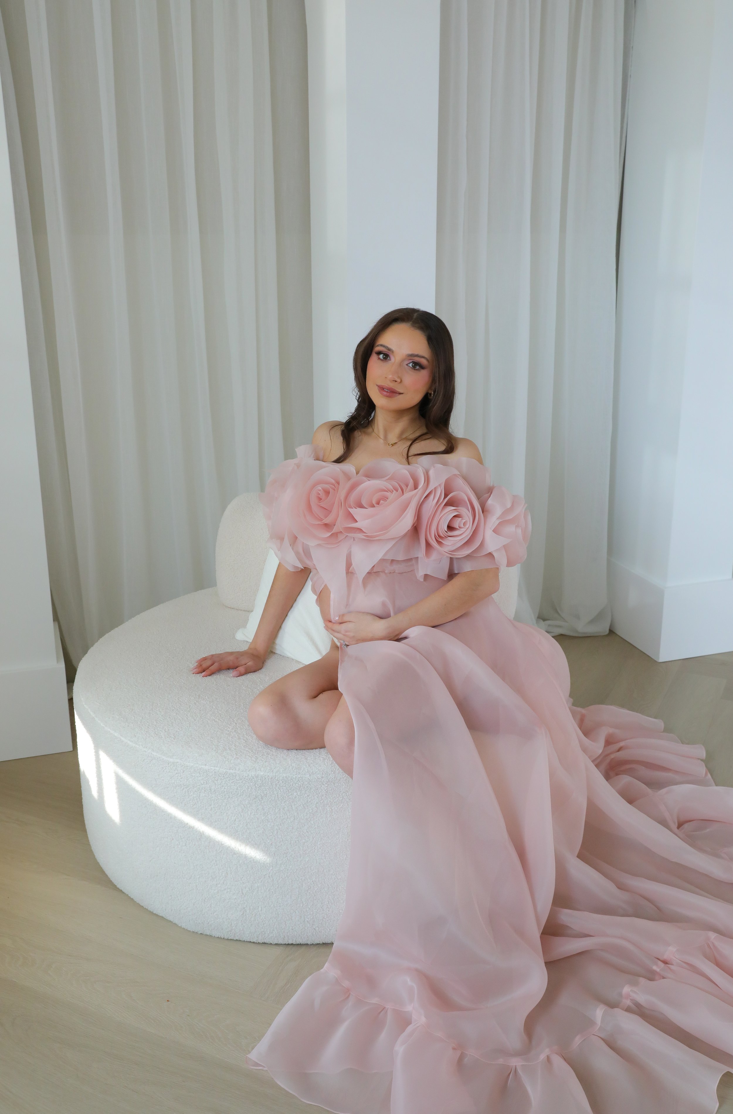A young woman in a pink gown with large fabric roses on the bodice, sitting on a white circular chair in front of white curtains.
