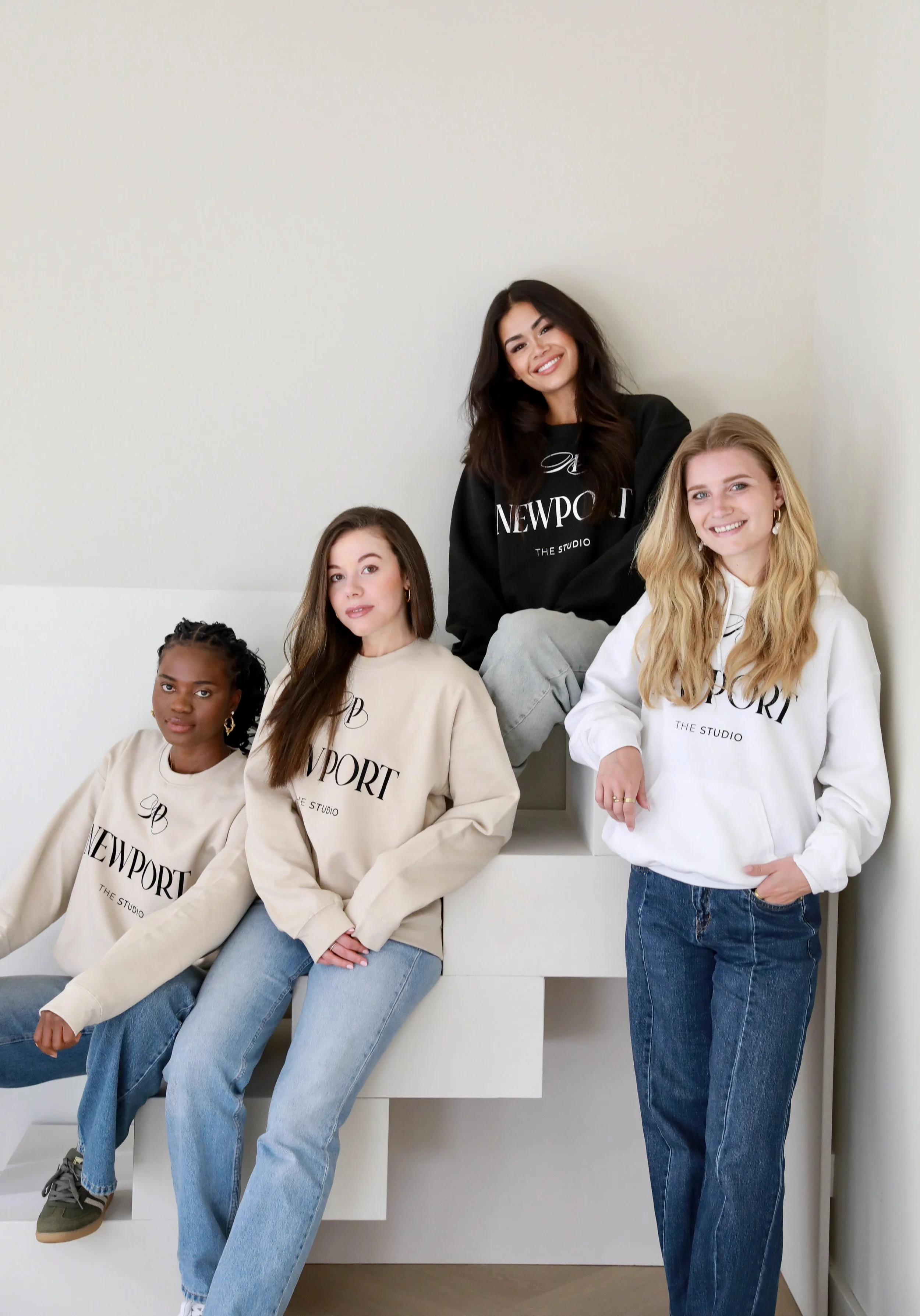 Group of five women standing and sitting on stairs, posing for a photo. They are wearing casual sweatshirts with the word 'NEWPORT' printed on them.