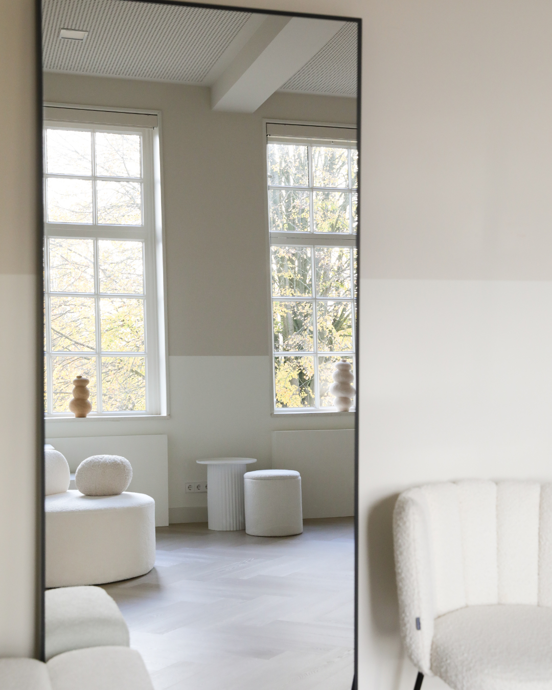 Minimalist white living room with large windows, white furniture, and decorative objects in neutral tones, seen through a mirror.