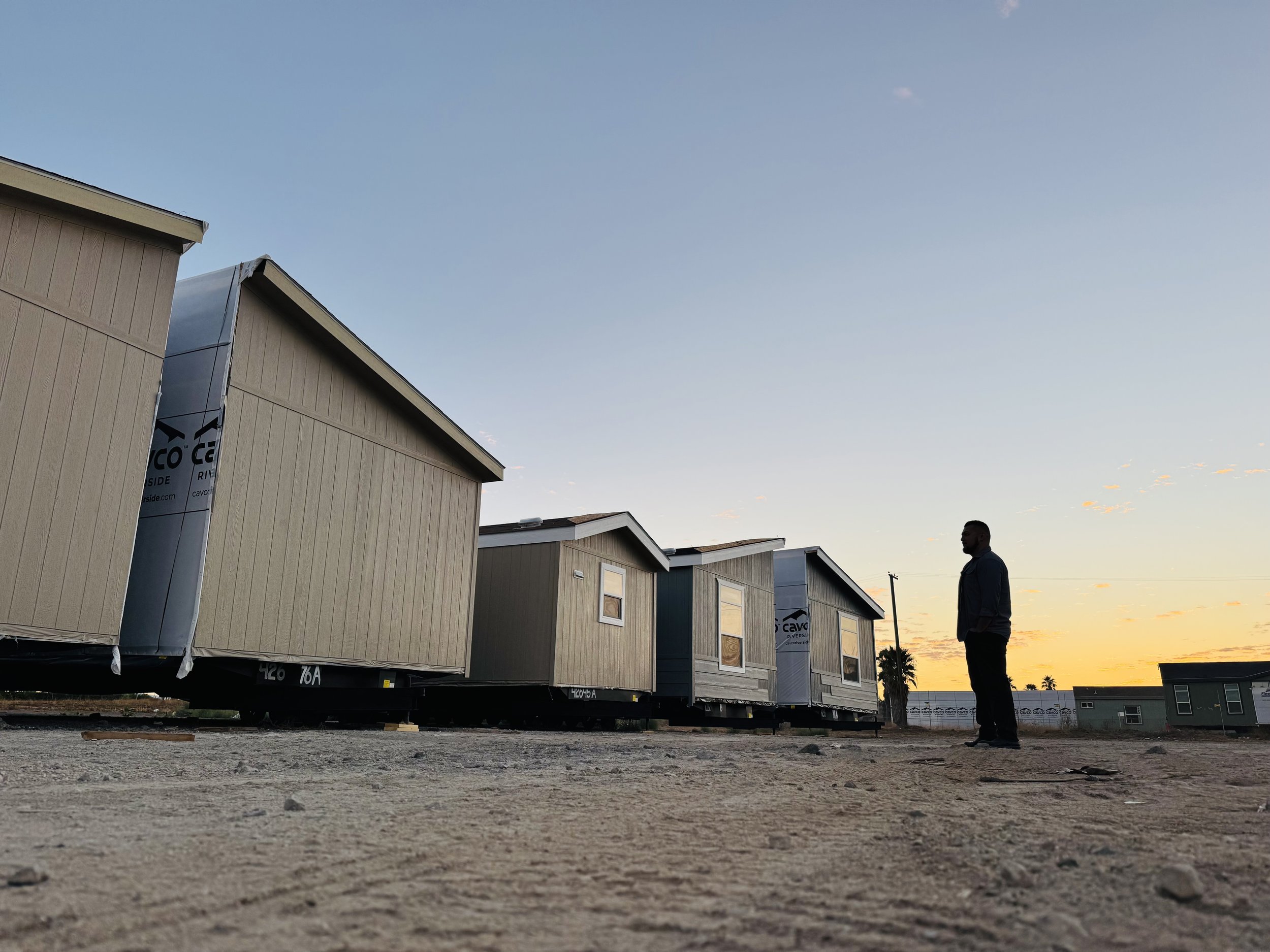 Housing units under construction at sunset, with a man standing nearby on a dirt ground.
