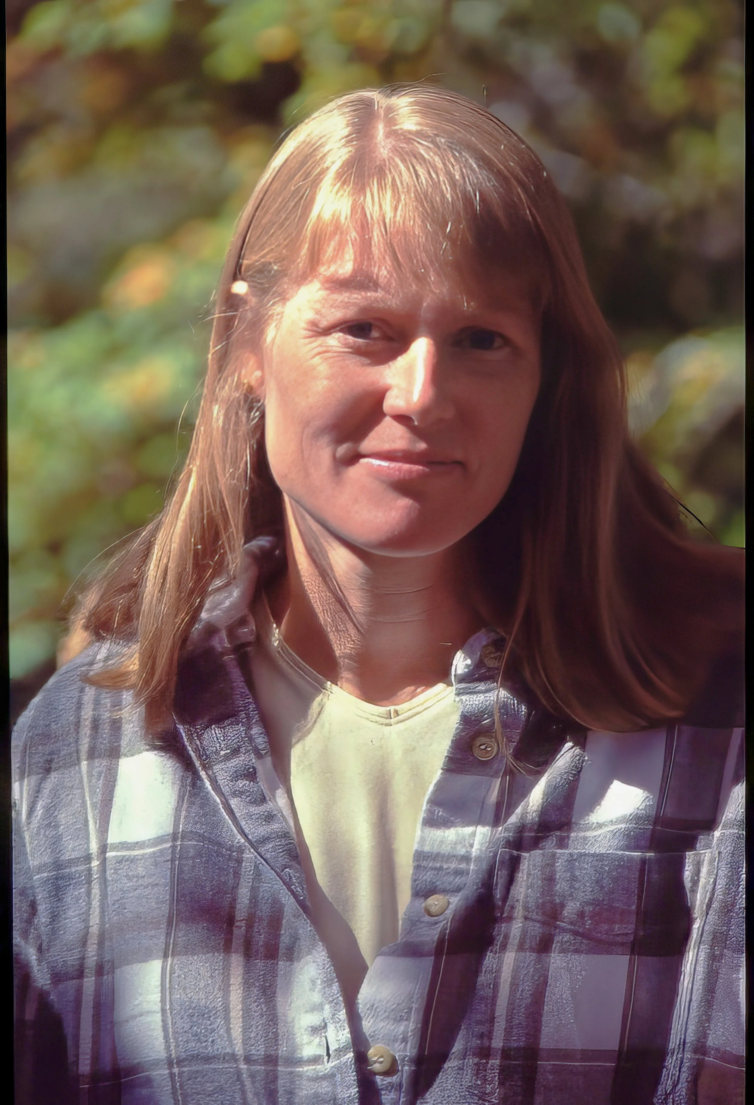 A woman with long brown hair wearing a plaid shirt, one button unbuttoned, standing outdoors with a blurred green and brown background.