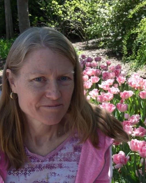 A woman with long blonde hair and blue eyes standing in front of a bed of pink tulips in a garden with green trees and bushes.