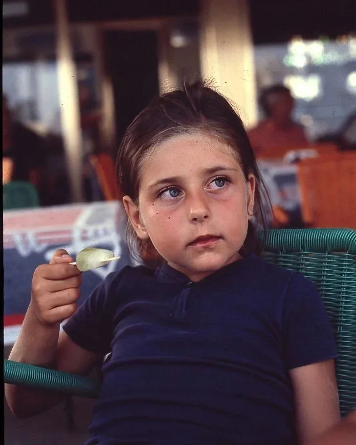 A young girl with light brown hair and blue eyes is sitting at a table in a restaurant or cafe. She is wearing a dark navy shirt and holding a tablespoonful of food, possibly pudding or ice cream, in her right hand. The background includes other patr