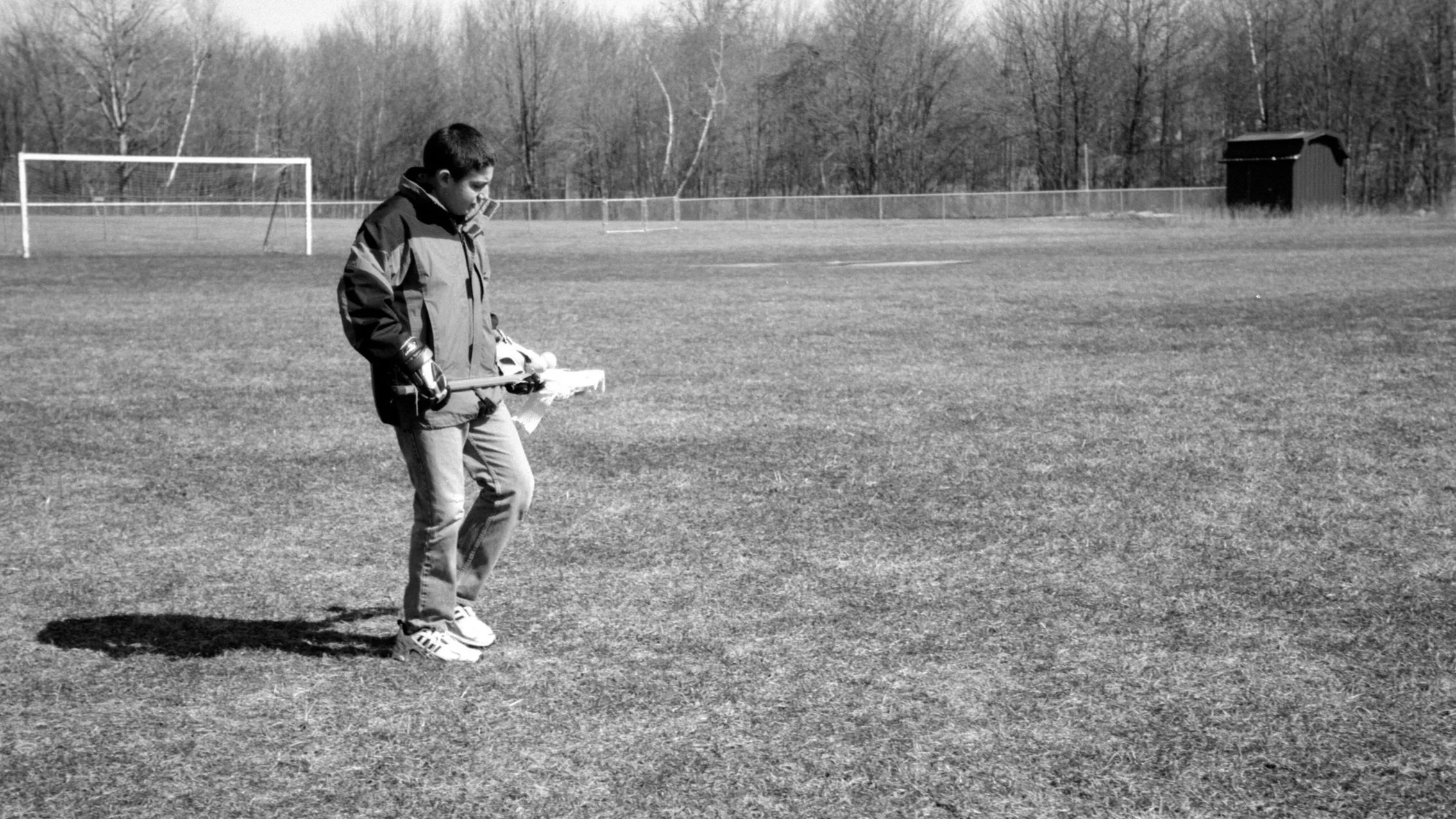 A boy in a jacket and jeans is standing on a grassy field holding a hockey stick, with a patch of shadow on the ground nearby. In the background, there are goalposts, a fence, trees, and a shed.