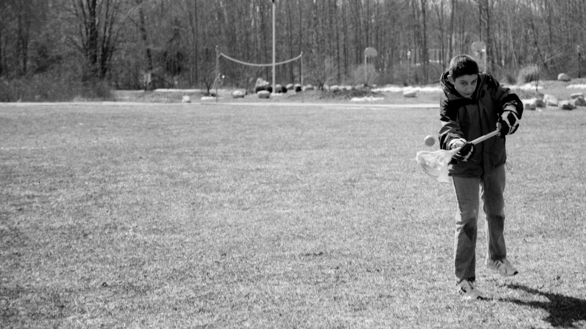 A boy playing lacrosse on an open grassy field, wearing a jacket and gloves, with trees and rocks in the background.