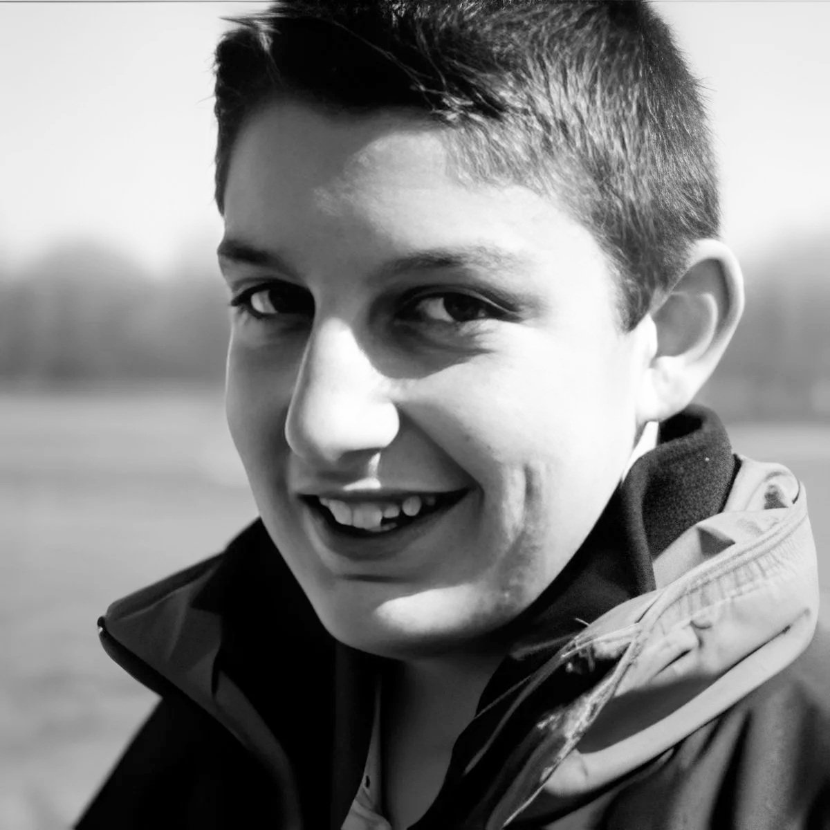 Close-up black-and-white photo of a smiling young person with short hair, wearing a jacket outdoors.