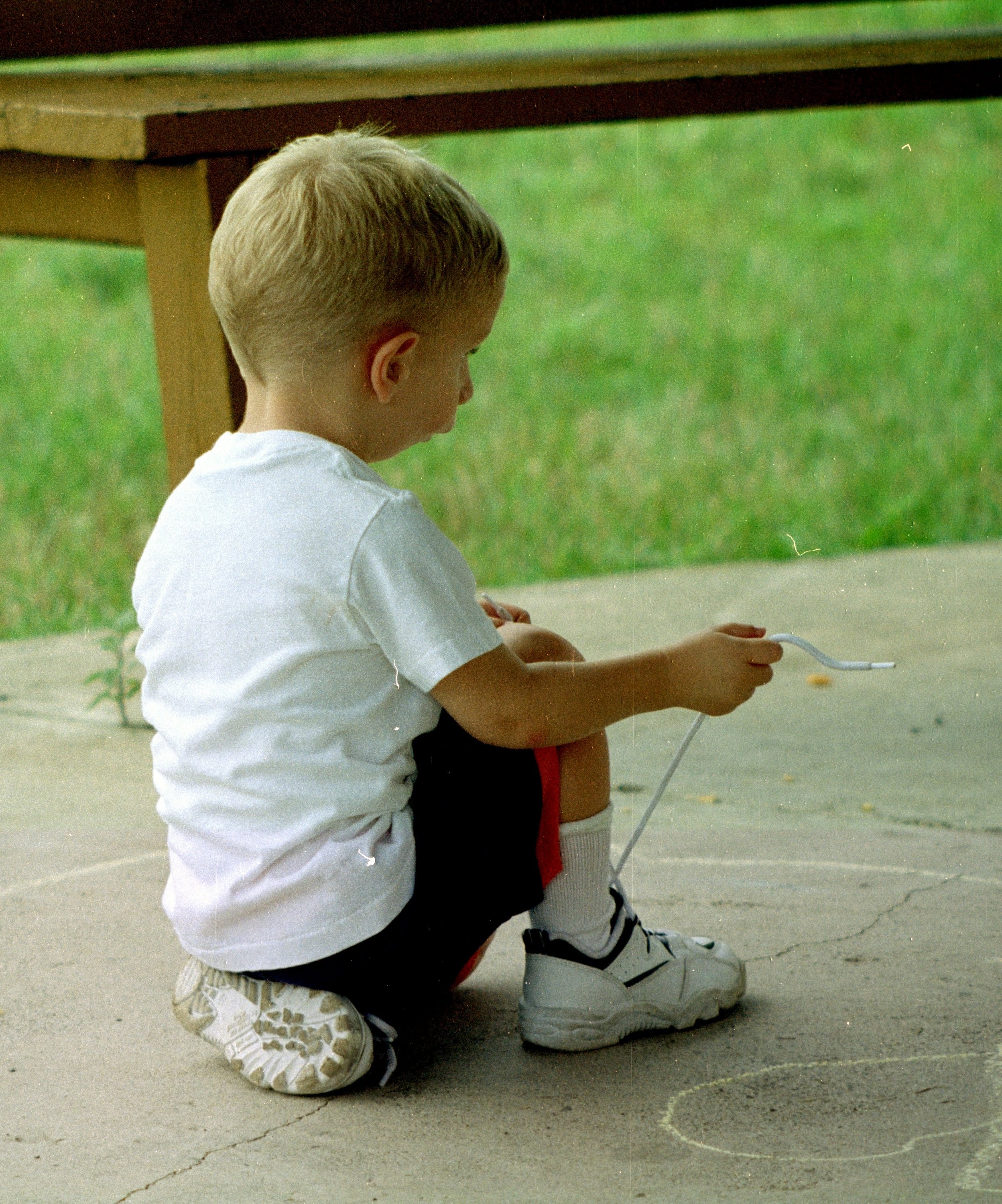 A young boy in a white t-shirt, black shorts, and white sneakers is squatting on a concrete sidewalk tying his shoe, drawing with chalk on the ground. There is a wooden picnic table in the background on a grassy area.