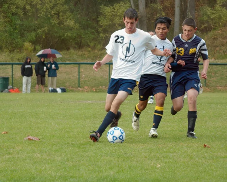 Three boys playing soccer on a grassy field with a fence and trees in the background. Two boys wearing white shirts and one in a striped blue shirt are competing for the soccer ball.