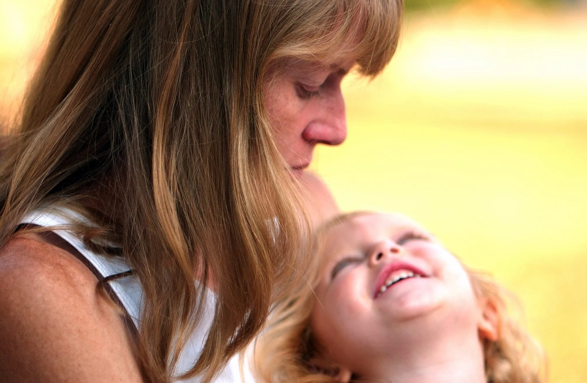 A woman and a young girl share a close, joyful moment outdoors, with the woman gently nuzzling the girl who is smiling and looking up at her.