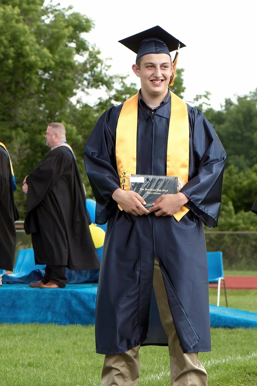 A young man in a blue graduation gown and cap with a gold stole is standing outdoors on a grassy field, holding a diploma case, and smiling. Other graduates are in the background.