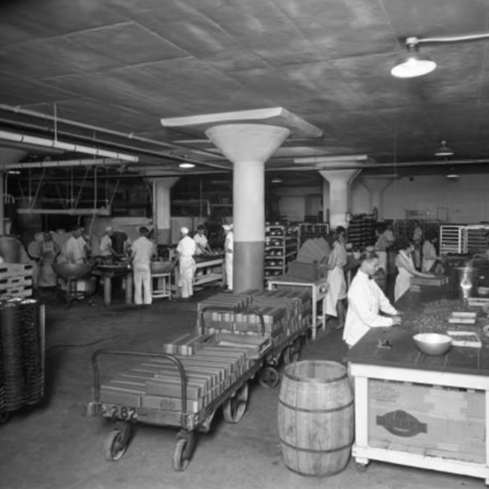 Workers in a large industrial kitchen or bakery preparing food with various tables, carts, and kitchen equipment.
