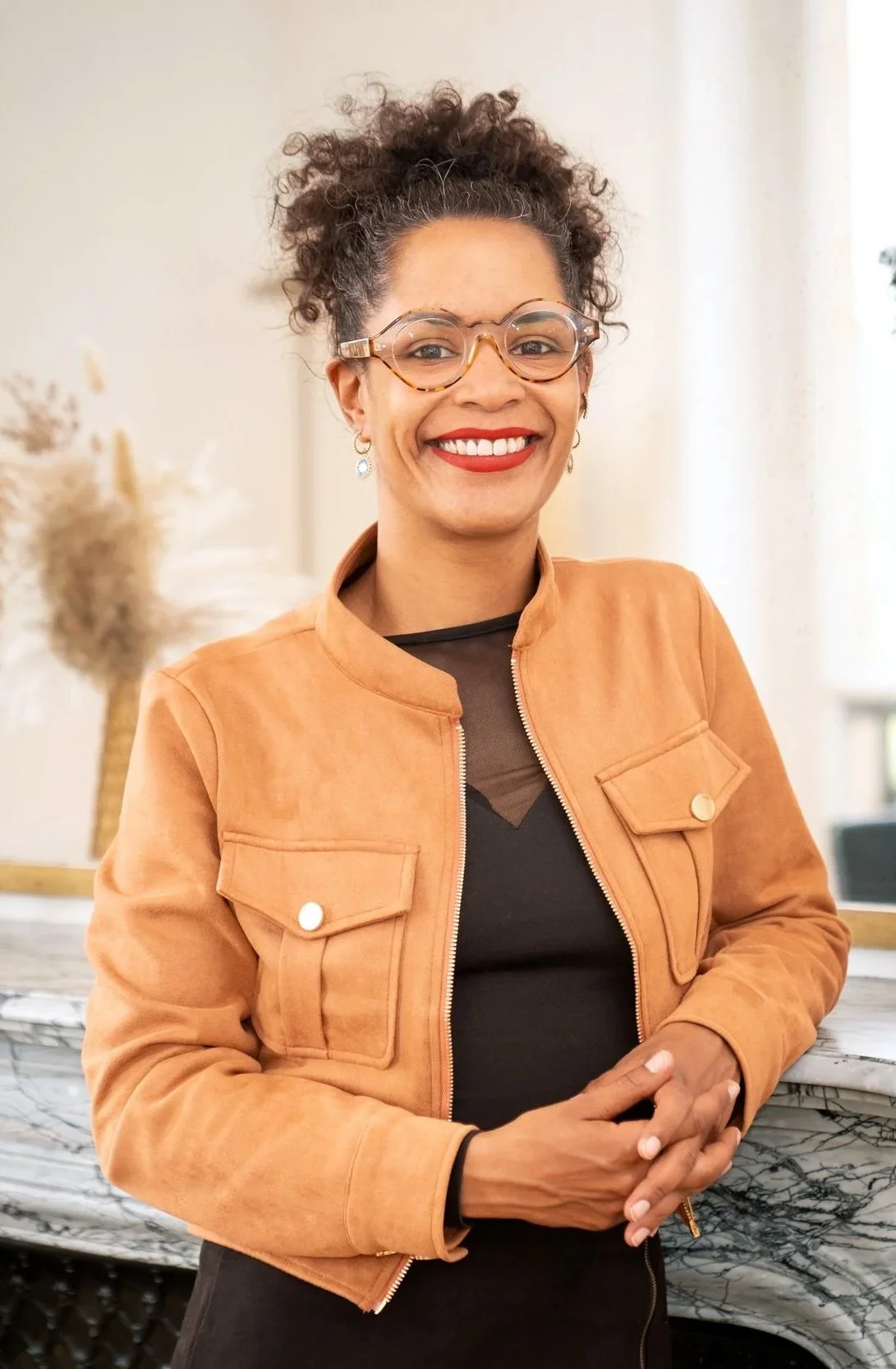 Une femme souriante portant des lunettes tendance, une veste en cuir orange et un haut noir, debout près d'un comptoir en marbre dans une pièce lumineuse.