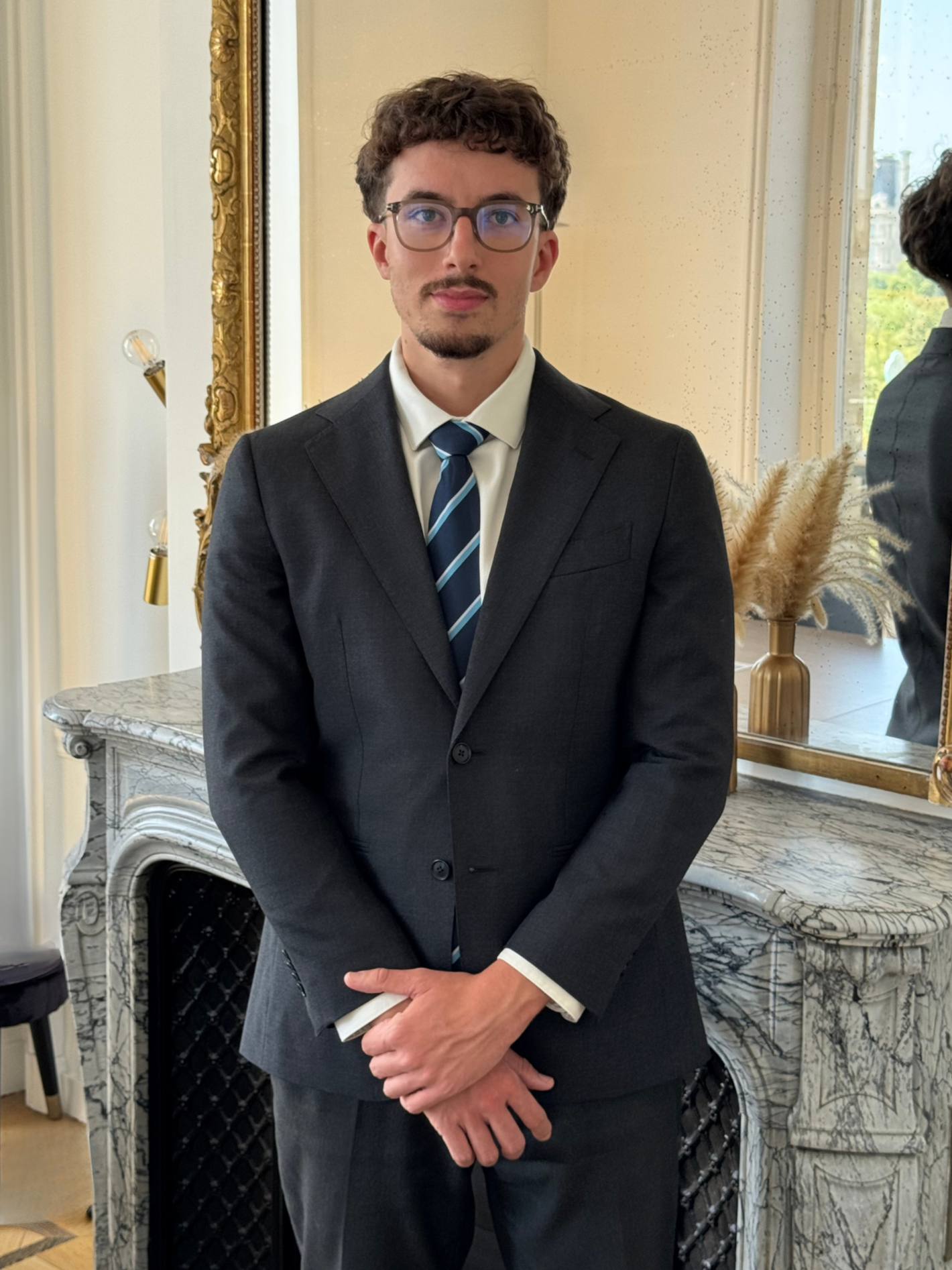 Un jeune homme en costume sombre, chemise blanche et cravate bleue, portant des lunettes, se tient dans une pièce élégante avec un miroir et un décor doré derrière lui.