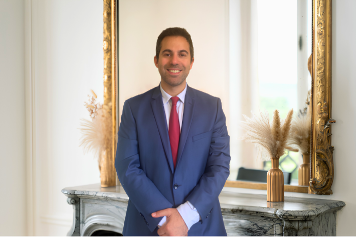Yoann Castiel, co-fondateur de Kays Wealth Management, spécialiste des produits structurés en costume bleu, cravate rouge, souriant, debout devant un miroir doré dans une pièce bien éclairée avec un décor élégant.