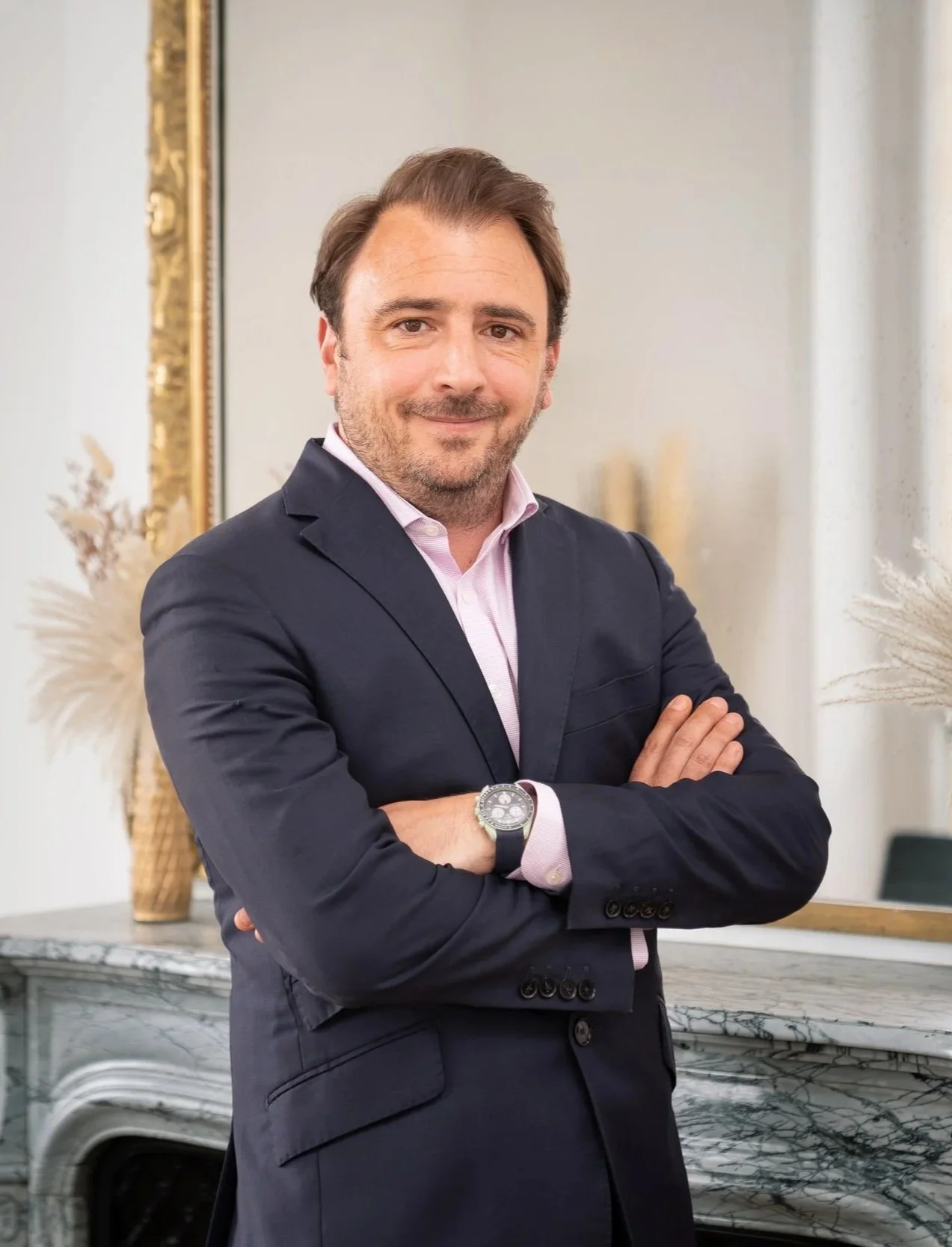 Homme d'affaires avec un costume sombre, coiffé et souriant, croisant les bras, dans un intérieur moderne avec un grand miroir doré, un mur blanc et une table en marbre.