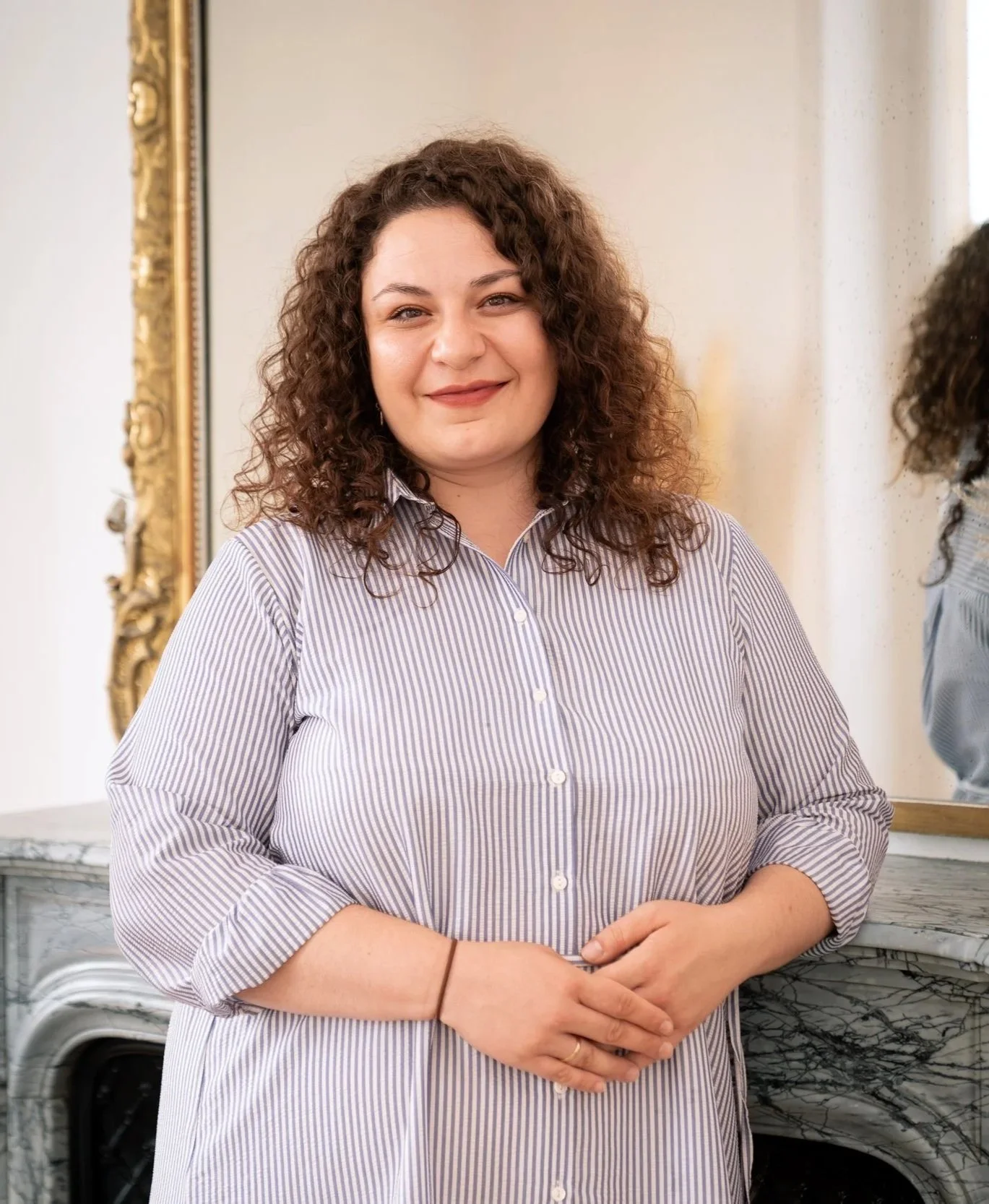Une femme avec des cheveux bouclés marron foncé portant une chemise à rayures. Elle se tient dans une pièce élégante, avec un miroir doré en arrière-plan et un mur blanc. Elle sourit légèrement.