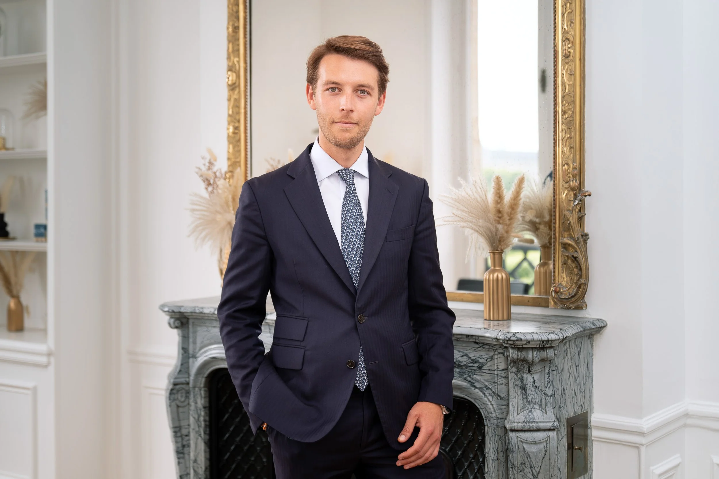 Homme en costume sombre, debout devant une cheminée en marbre avec un grand miroir doré derrière lui.