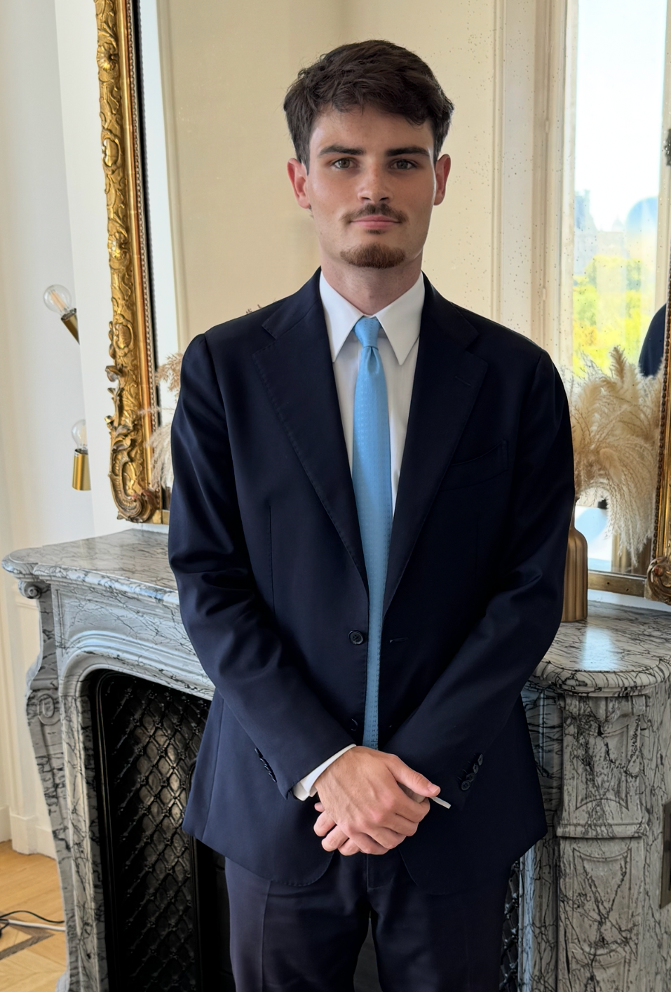 Jeune homme en costume noir avec une cravate bleue, posant dans une pièce élégante avec un miroir orné et une cheminée en marbre.