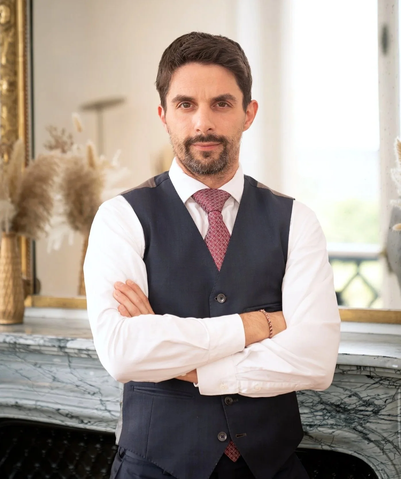 Un homme en costume costume avec une veste noire, une chemise blanche et une cravate rouge, croisant les bras, dans un intérieur avec un comptoir en marbre et des décorations de fleurs.
