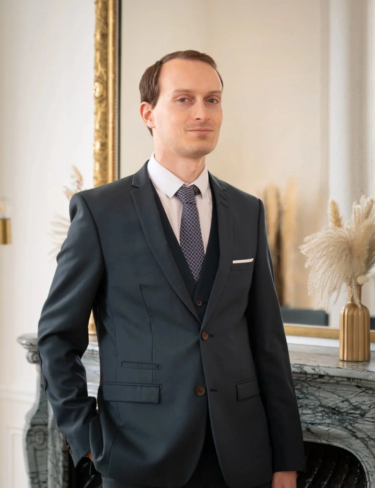 Un homme en costume noir, debout dans une pièce élégante avec un miroir et une console en marbre avec une plante décorative.