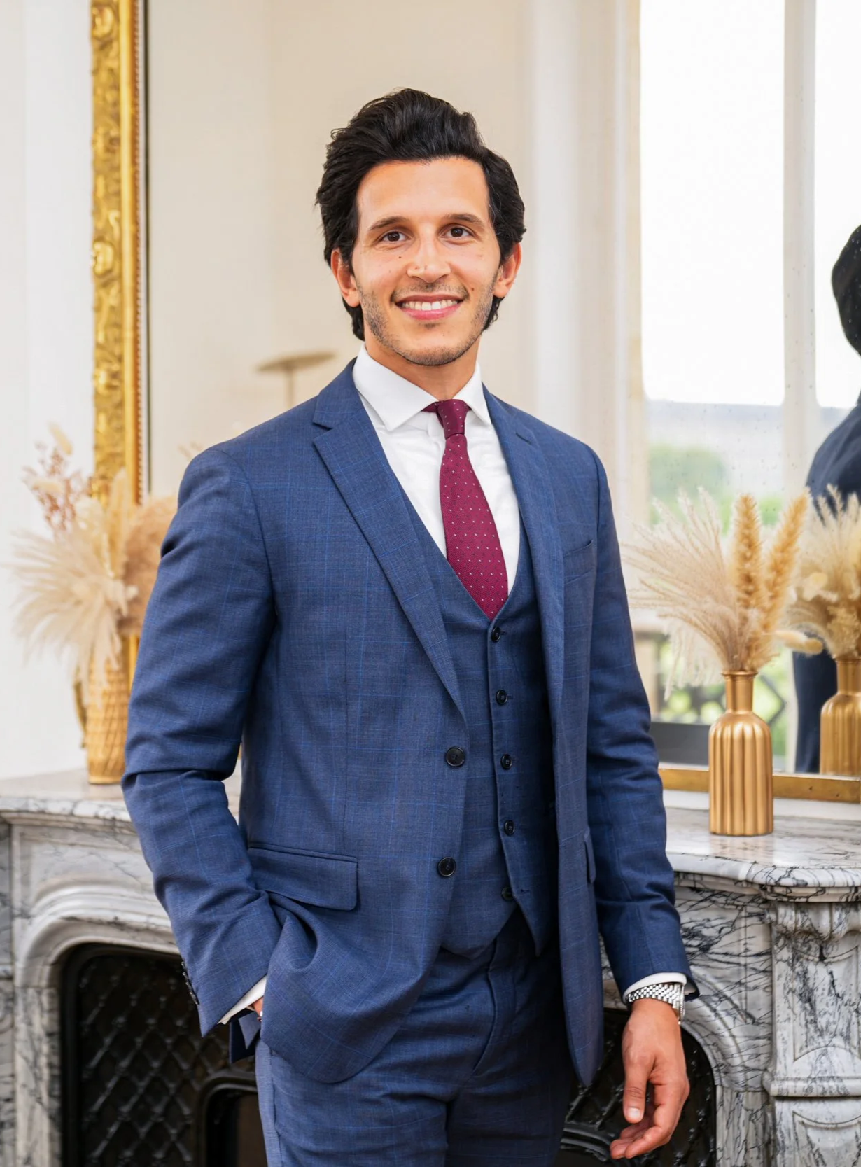 Mickaël Sahnoun, PDG de Kays Wealth Management souriant portant un costume bleu, une chemise blanche et une cravate rouge, debout dans un intérieur élégant avec un miroir, des vases avec des fleurs séchées et une cheminée en marbre.