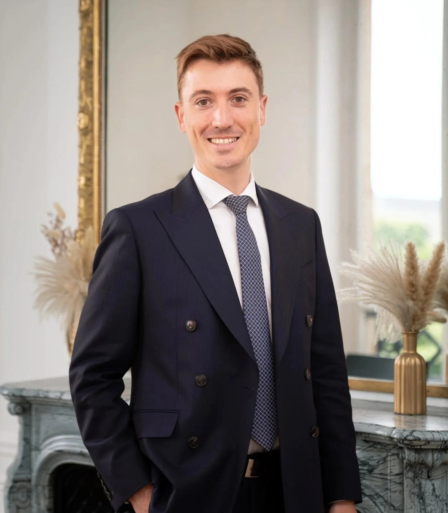 Un homme souriant, portant un costume noir avec une chemise blanche et une cravate, debout dans une pièce élégante avec un miroir doré, un vase en or et décorations florales.