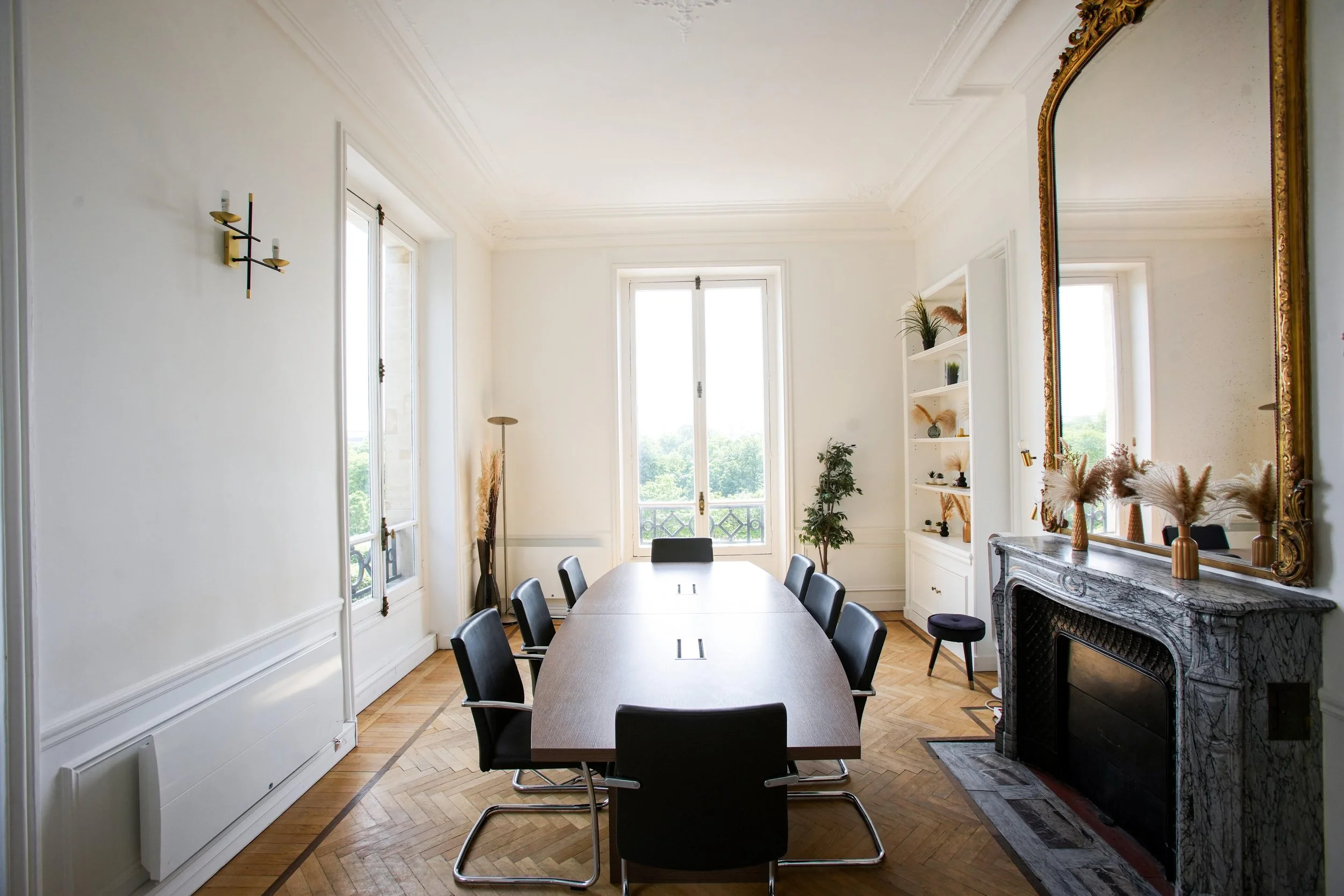 Salle de réunion spacieuse des bureaux Kays Wealth Management à Paris, rue de rivoli avec table, chaises, cheminée en marbre, grands fenêtres laissant entrer la lumière, étagères avec plantes et objets décoratifs, miroir ancien en cadre doré.