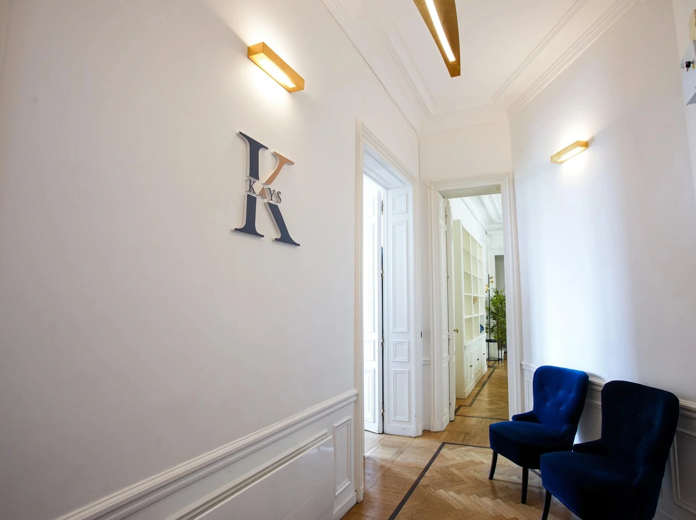 l'entrée lumineuse des ureaux de KAYS Wealth Mangement rue de Rivoli à Paris avec des murs blancs, un miroir mural avec le mot 'KEYS', des portes ouvertes donnant sur un couloir, fauteuil bleu et plantes.