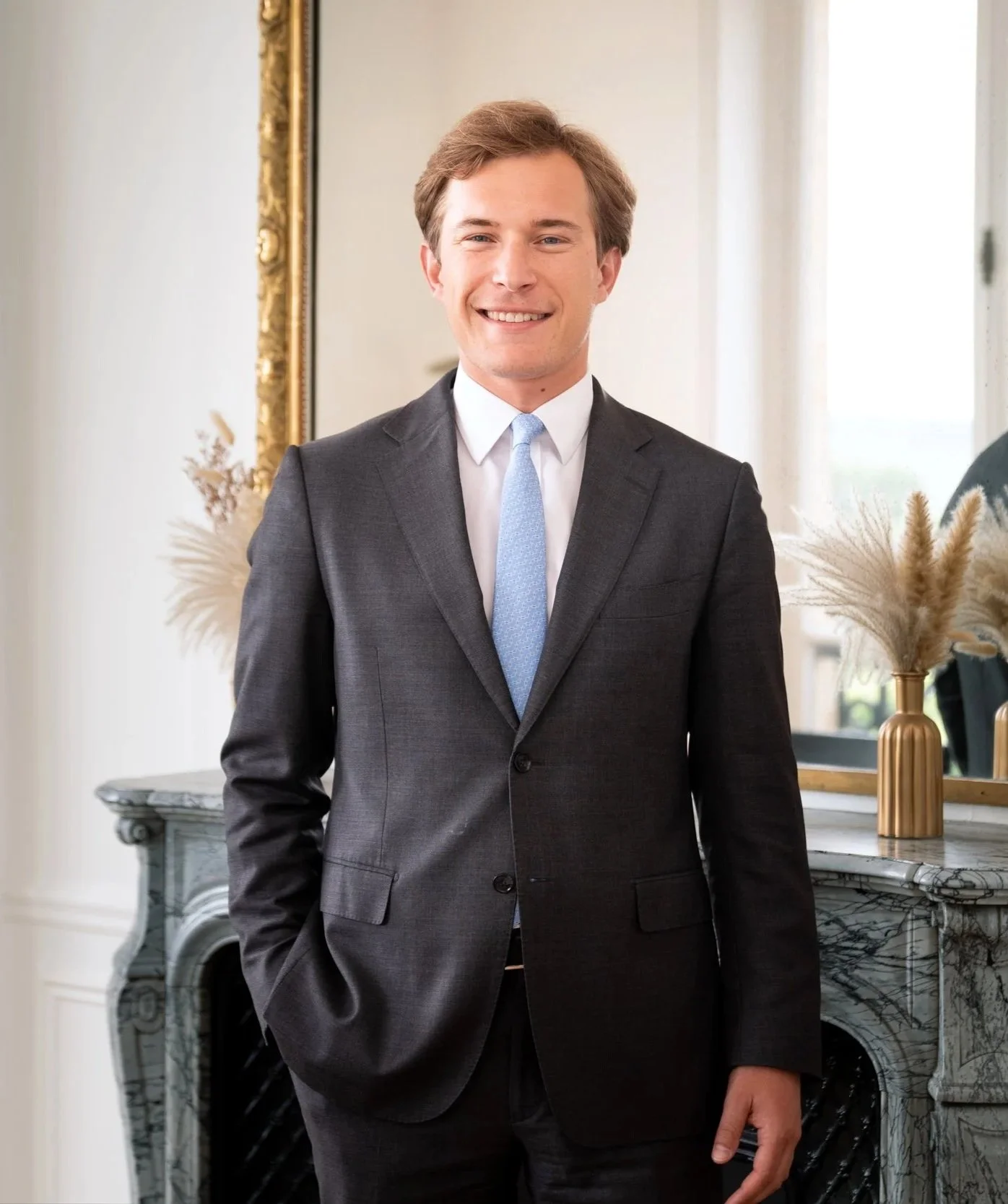 Jeune homme souriant en costume noir avec cravate bleue dans un intérieur élégant, devant une cheminée en marbre et un grand miroir doré.