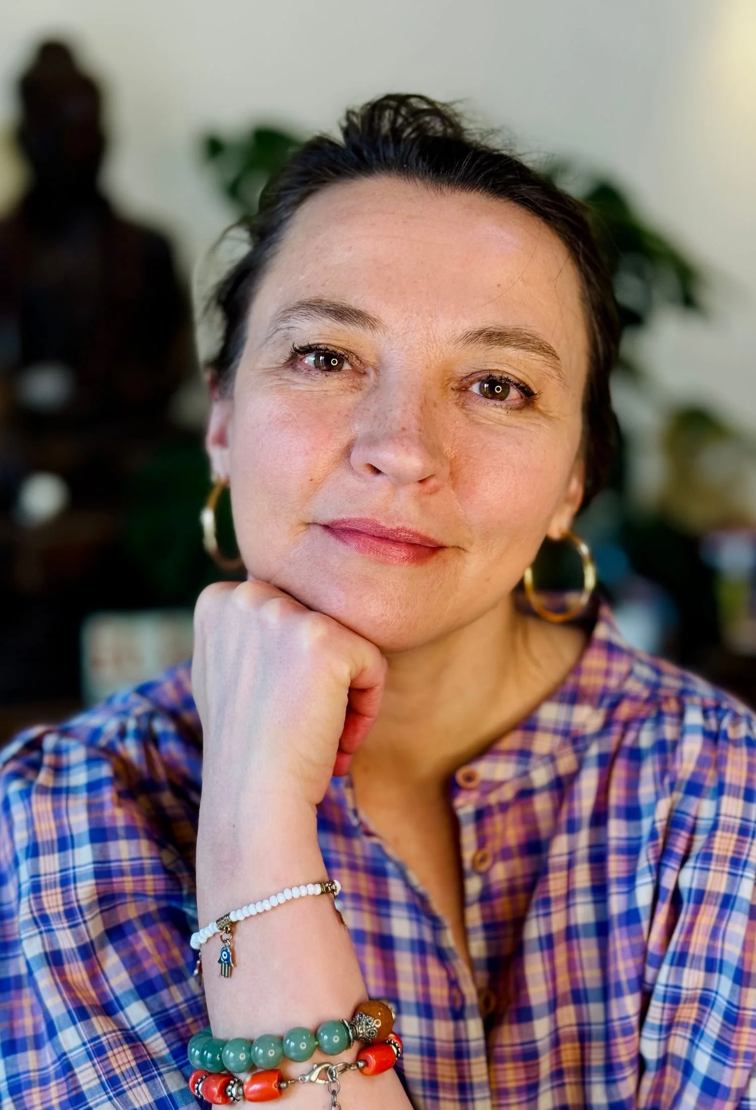 Close-up of a woman with dark hair, wearing a plaid shirt, gold hoop earrings, and colorful beaded bracelets, resting her chin on her hand with a blurred background featuring a statue and green plants.