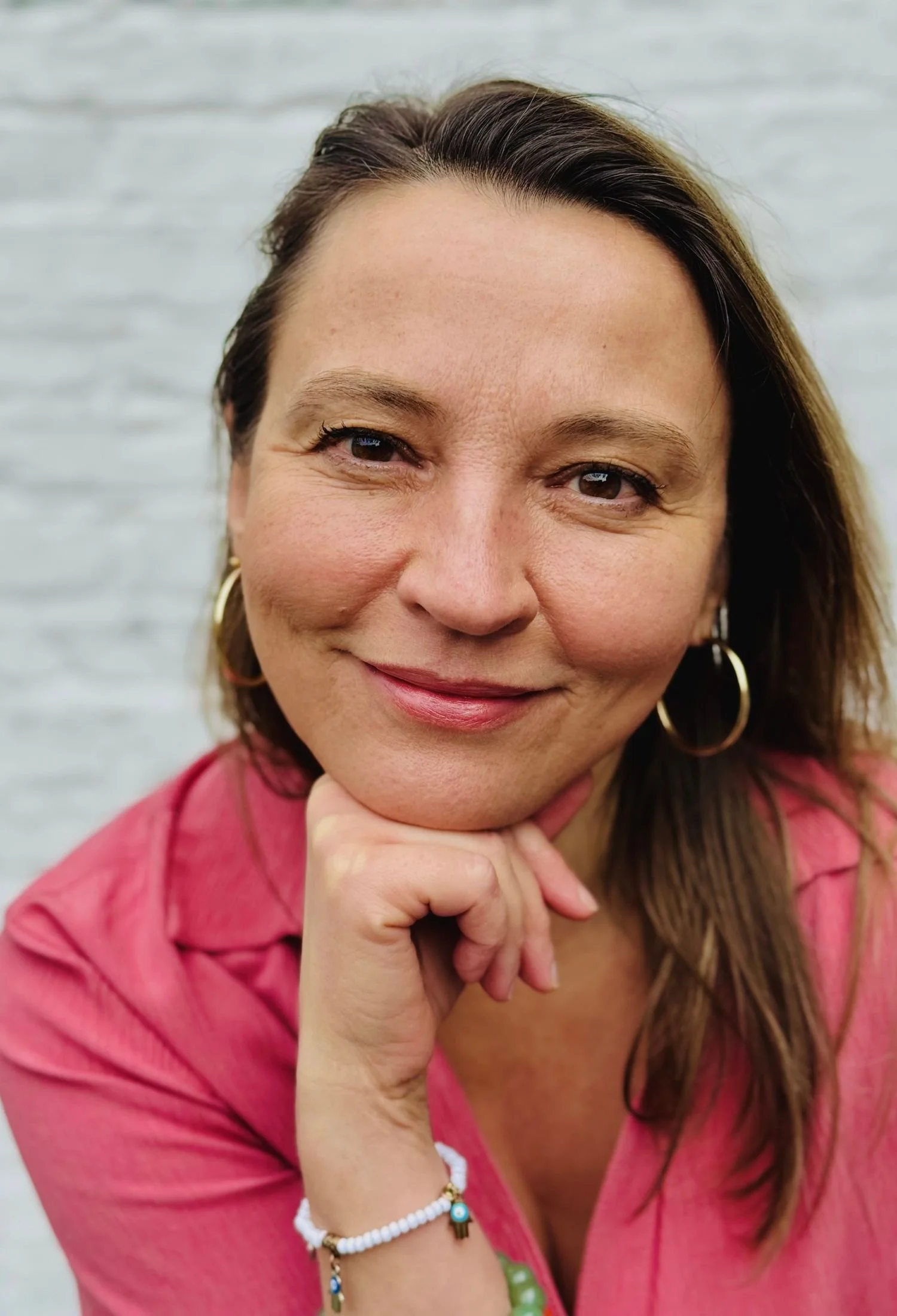 Close-up of a smiling woman with brown hair, wearing gold hoop earrings, a pink shirt, and bracelets, resting her chin on her hand in front of a white brick wall.