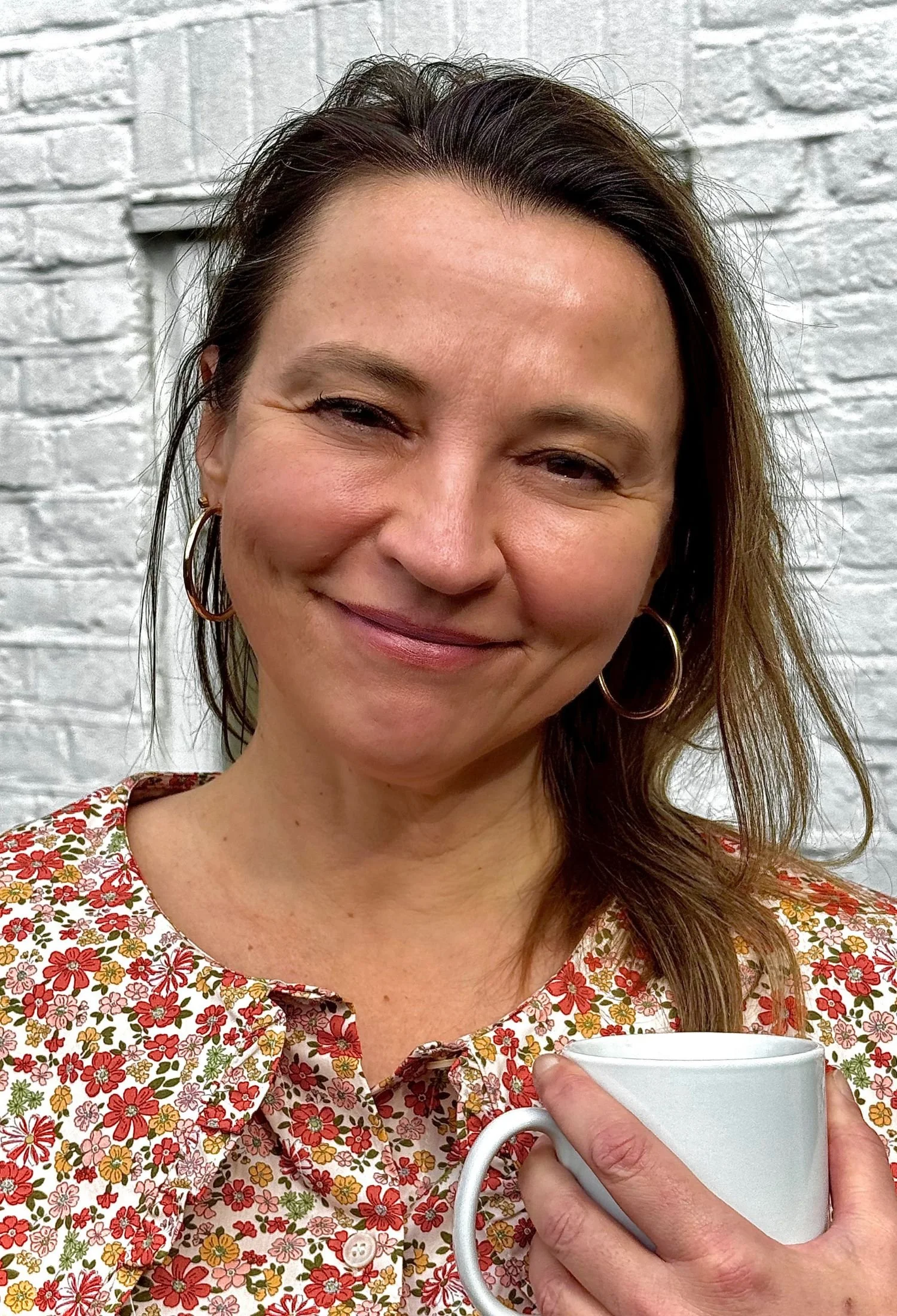 A woman smiling while holding a white mug against a white brick wall background. She is wearing a floral blouse and hoop earrings.