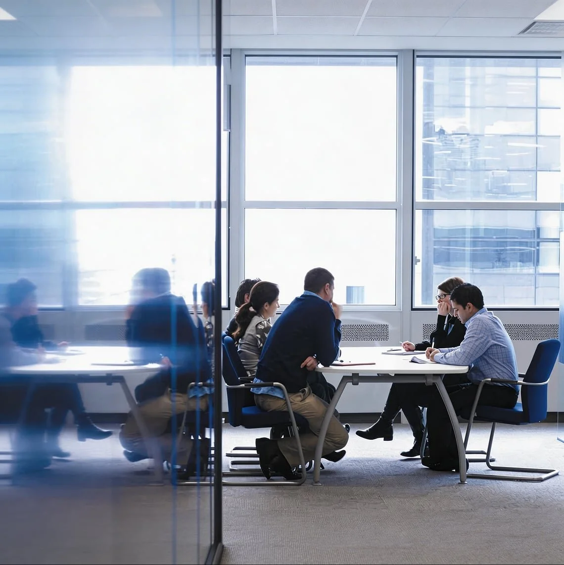 Business meeting in a modern conference room with large windows, featuring six people sitting around a table, engaged in discussion.