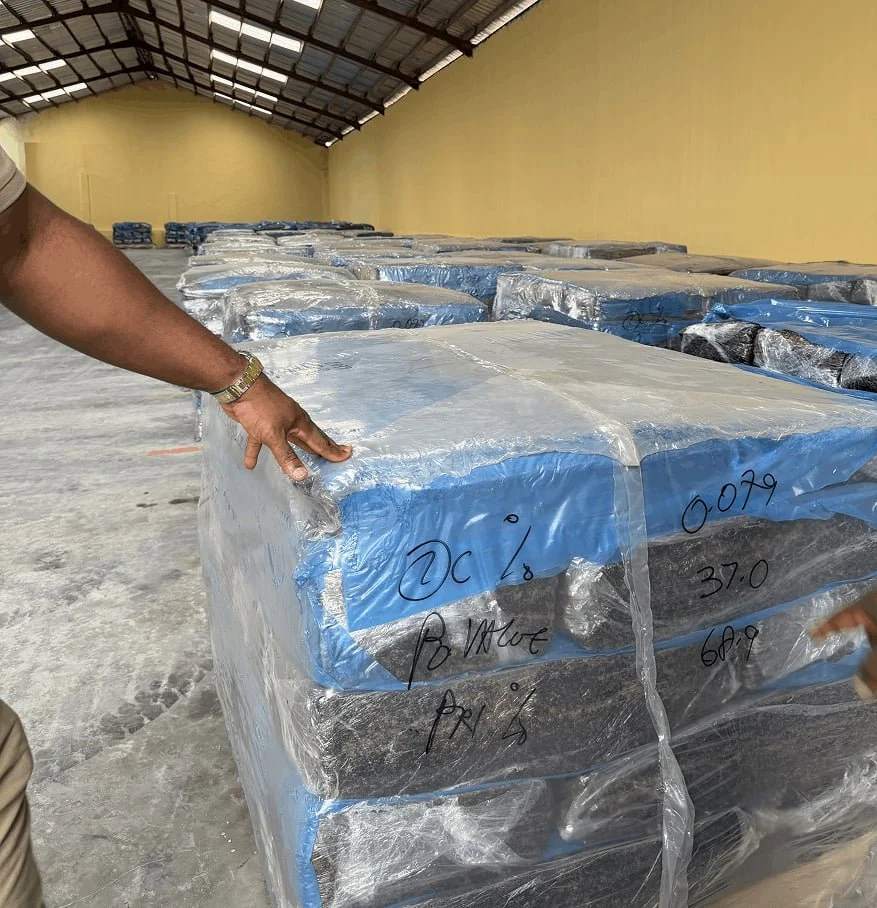 Large pallet wrapped in clear plastic containing blocks labeled with handwritten information, stored in a spacious warehouse with a high metal roof and yellow walls.
