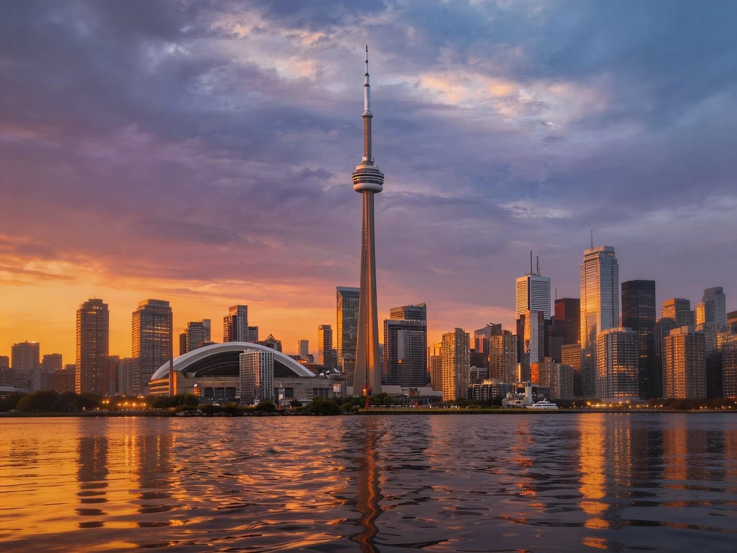 Sunset over the Toronto skyline with the CN Tower in the center, surrounded by modern skyscrapers reflected on Lake Ontario.