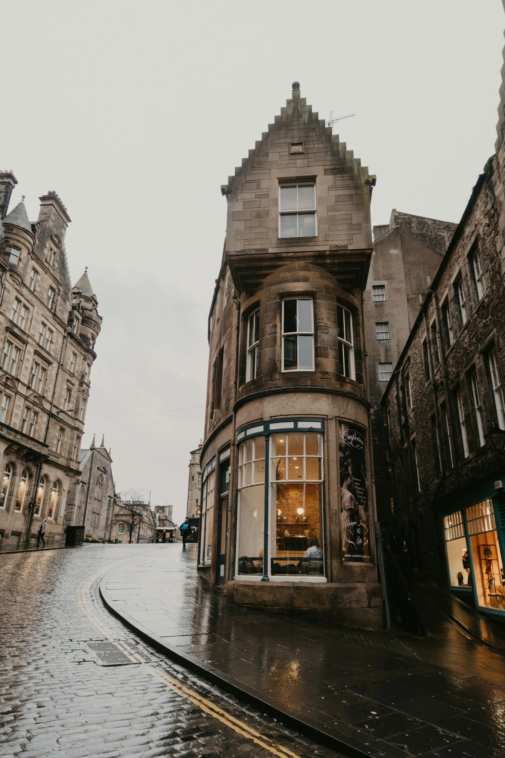 A narrow, cobblestone street with wet surface, curving to the left, lined with old historic buildings. The main building in focus is a corner structure with large display windows on the ground floor, a curved section, and a peaked roof. The street appears to be in a historic city center on a rainy day.