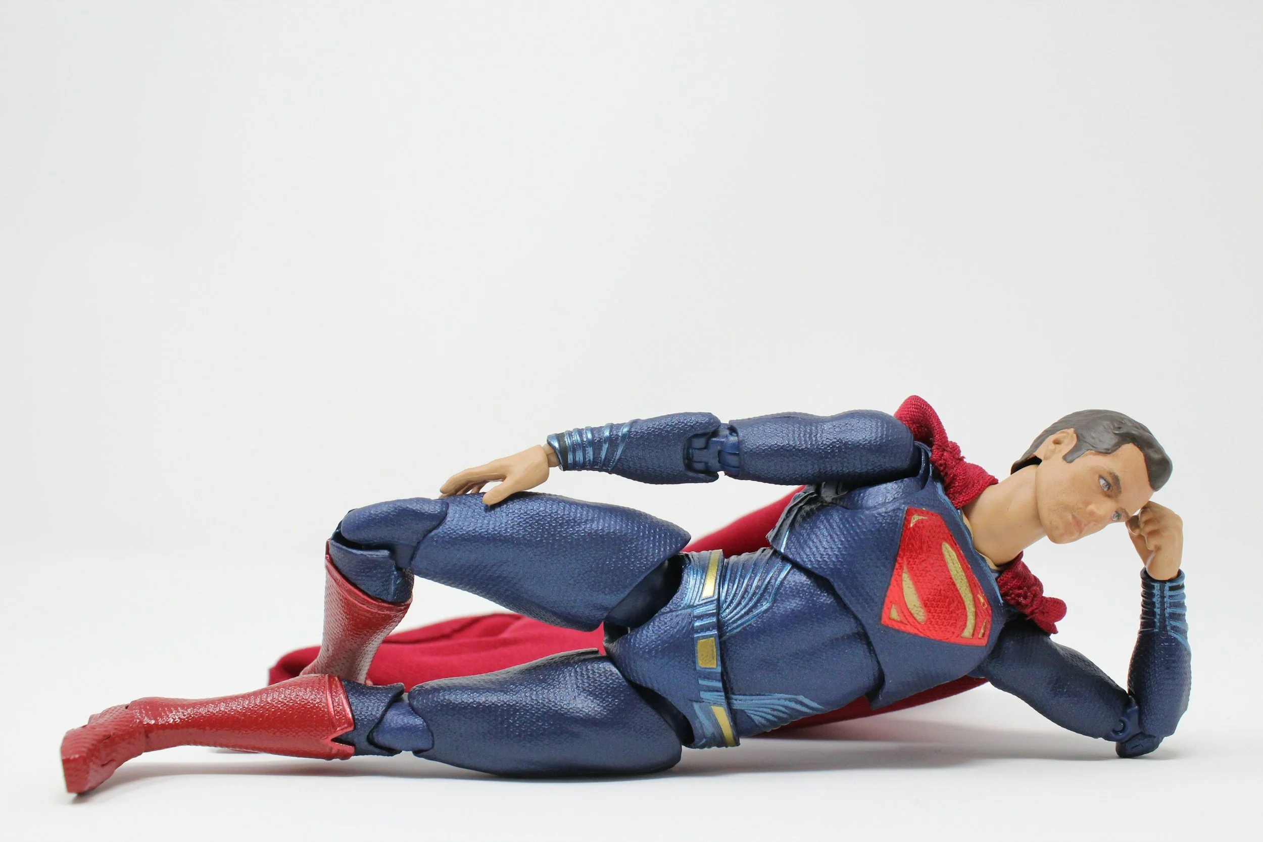 Action figure of Superman dressed in his iconic blue suit with red cape, red boots, and the Superman logo on his chest, lying on the ground in a replicated heroic pose.