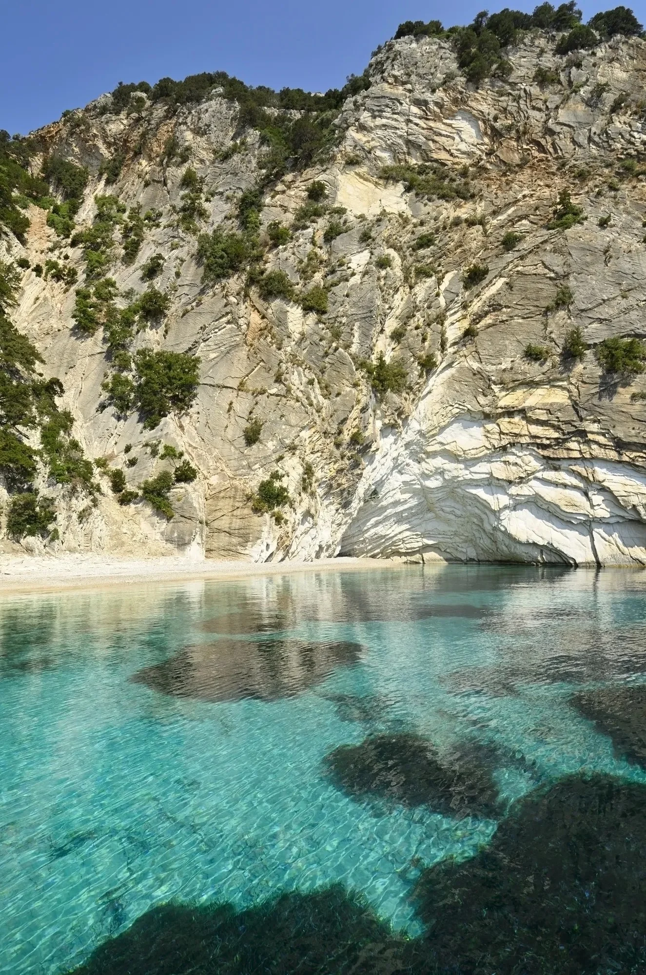 Cliffs with sparse vegetation falling into a clear turquoise body of water, reflecting the cliffs and the sky.