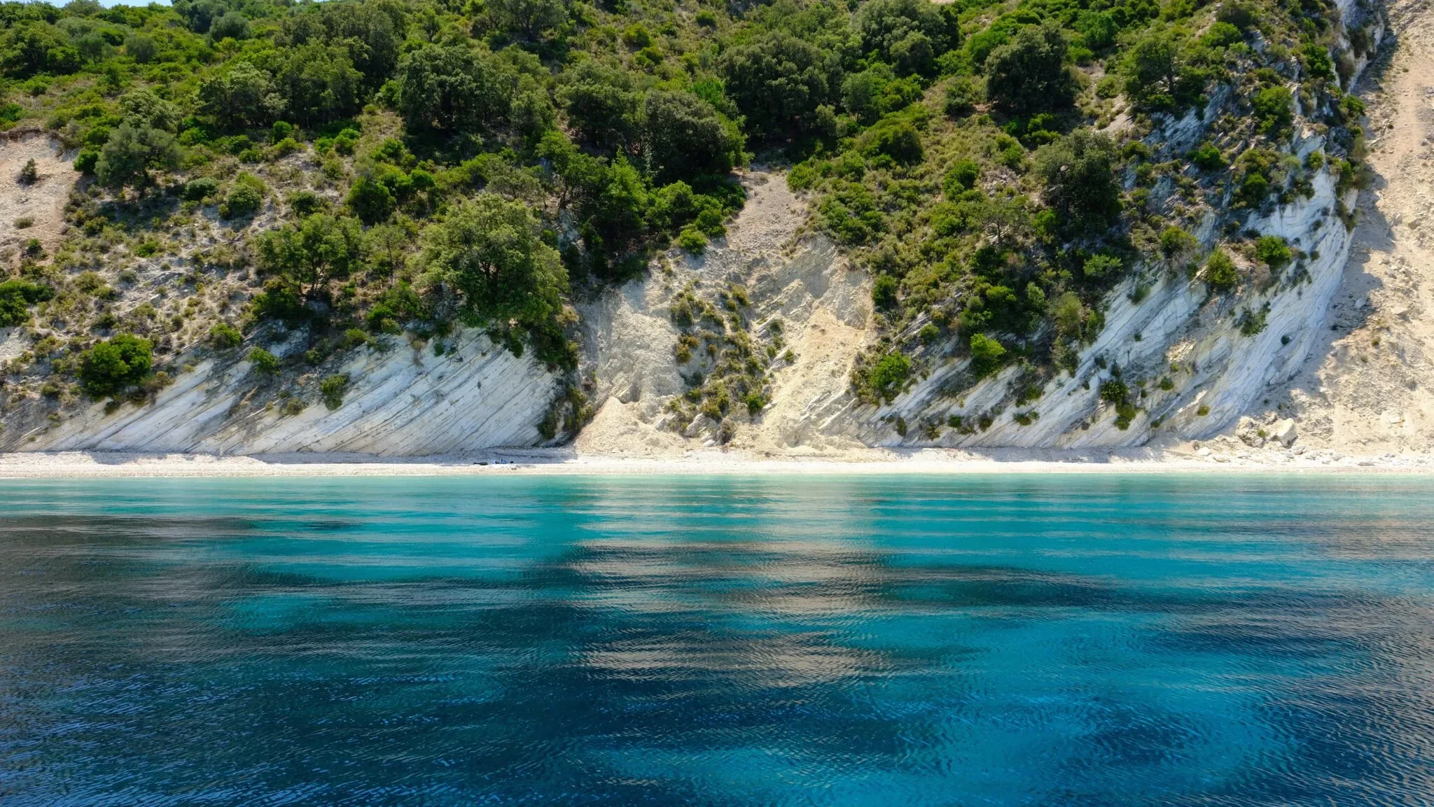 A large white chalk cliff with green trees and shrubs on top, adjacent to turquoise water with a reflection of the cliff and trees.