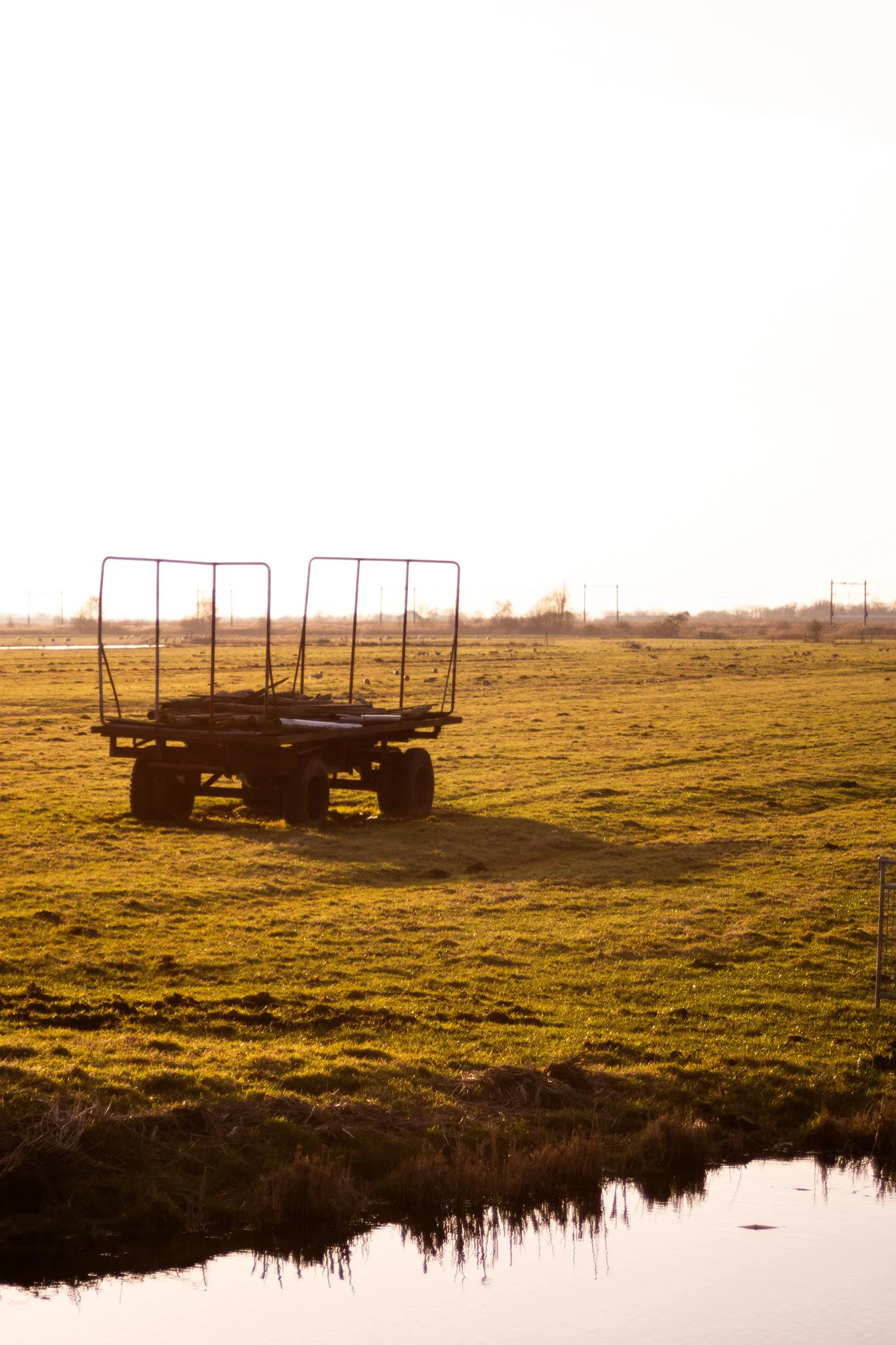 A small empty mobile golf cart or utility vehicle parked on a grassy field with a small pond in the foreground and power lines in the background, during sunset or sunrise.