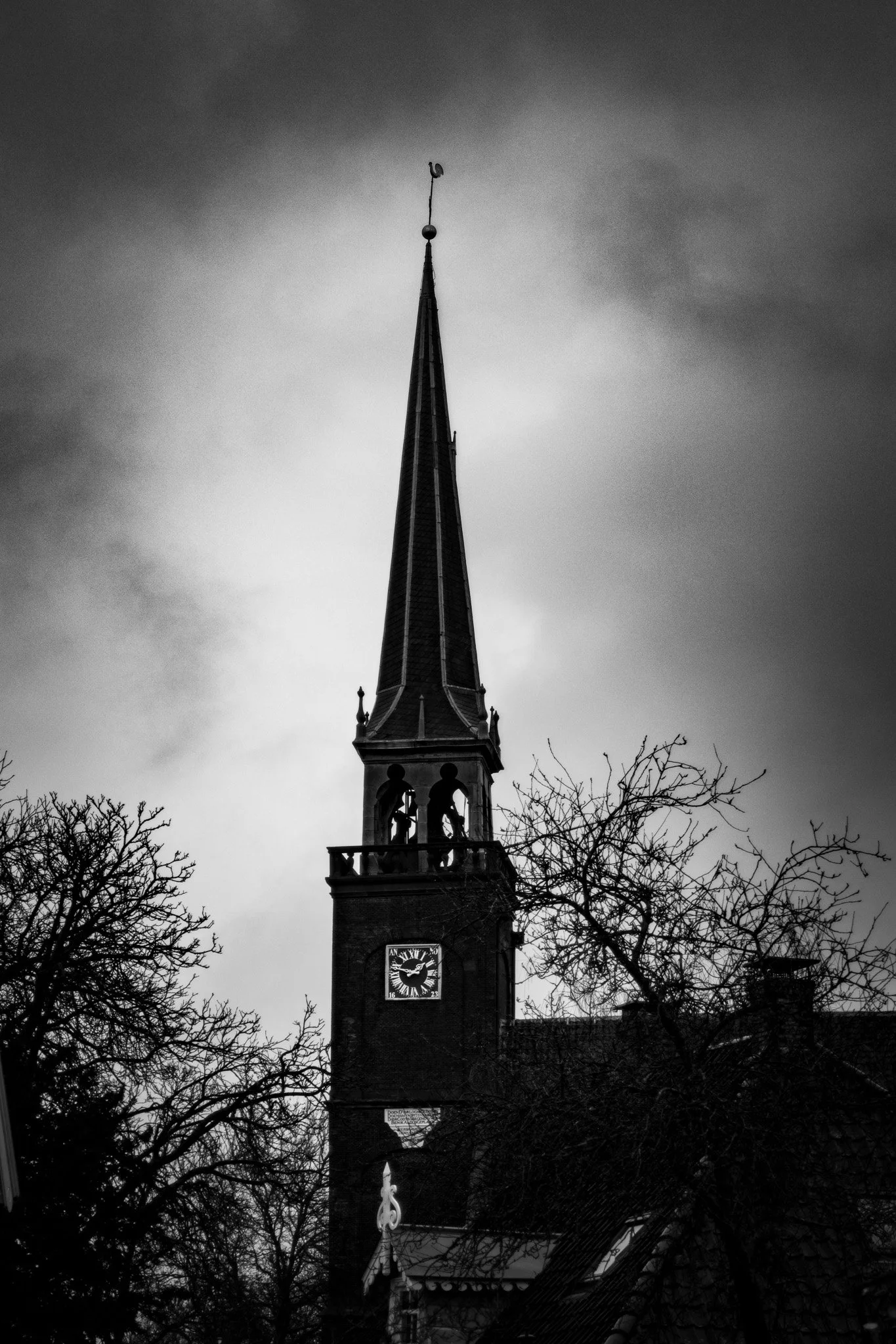 Black and white photo of a church steeple with clock, trees in foreground, cloudy sky in background.