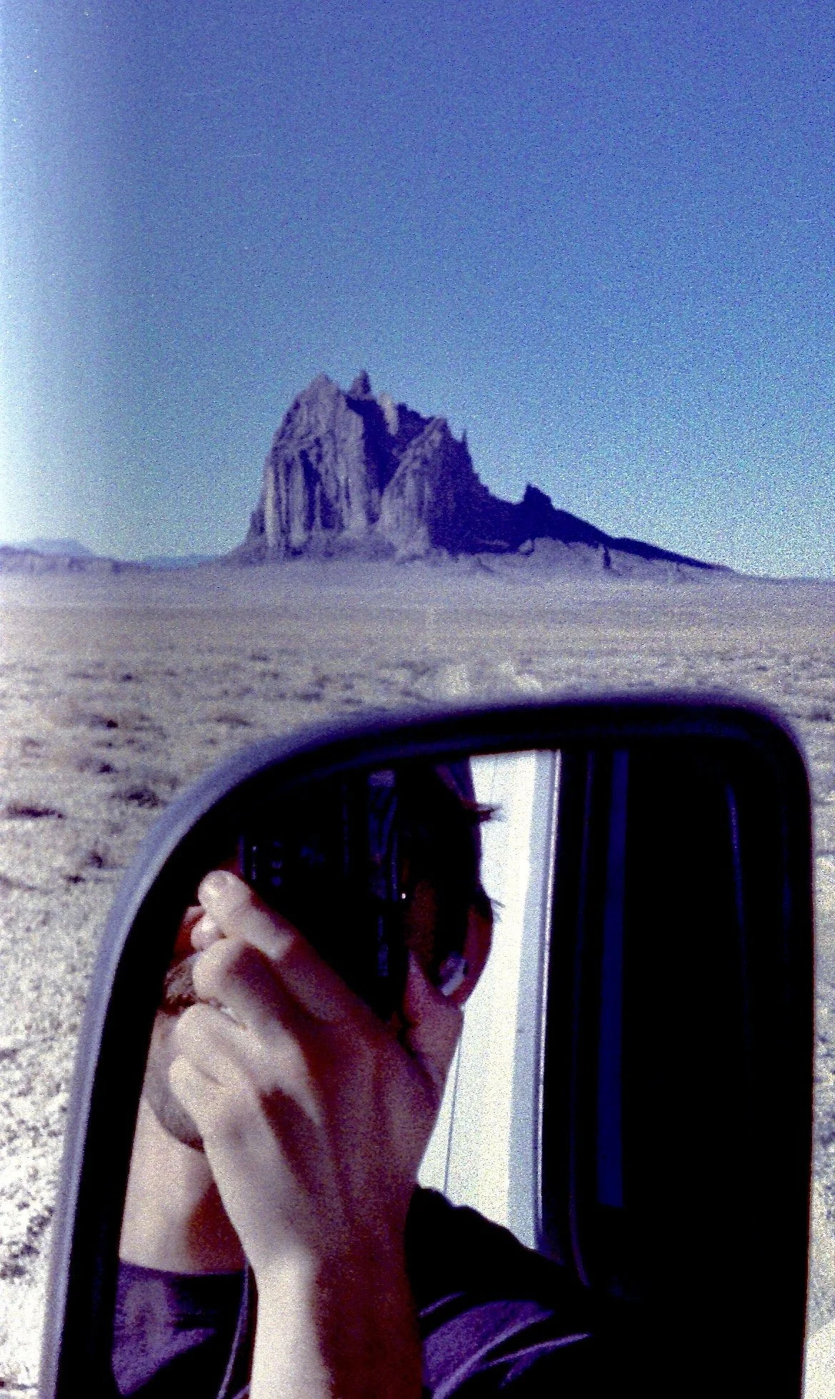 A person taking a selfie with a mountain in the background, seen in the side mirror of a vehicle.