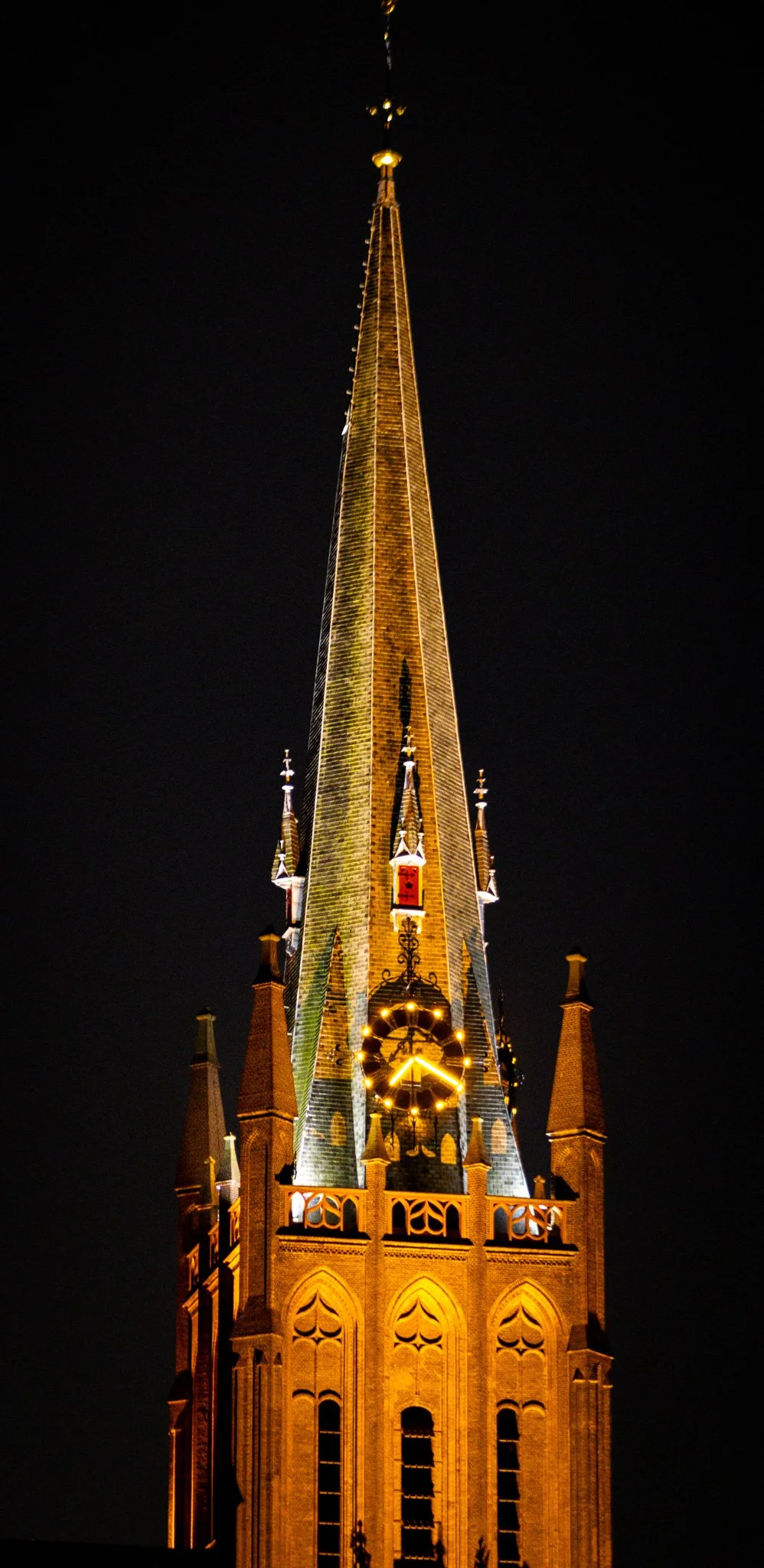 Night view of a tall, illuminated Gothic-style church steeple with a clock at the base, against a dark sky.