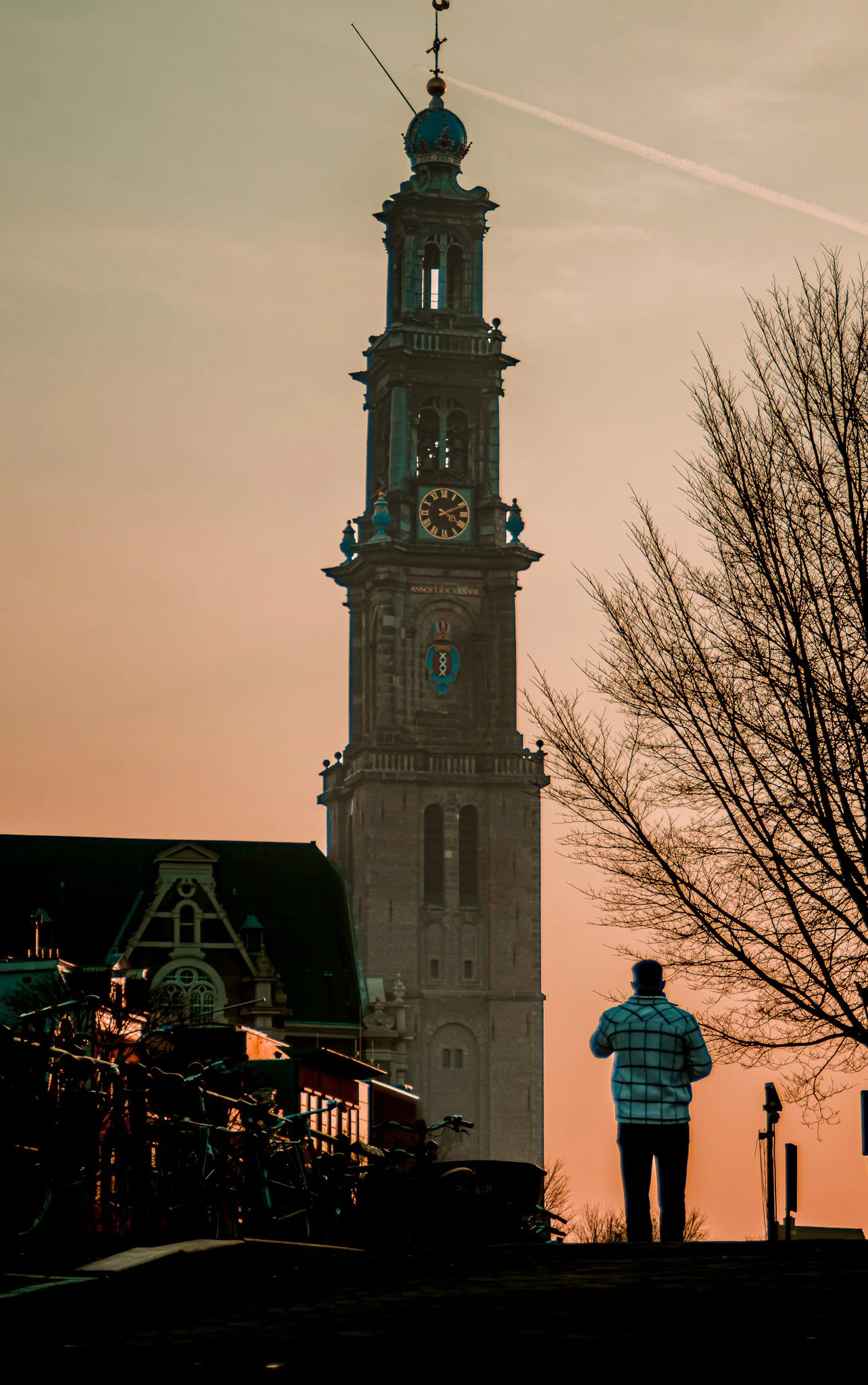 A person stands in front of a tall clock tower at sunset, with bicycles parked on the side and a leafless tree in the foreground.